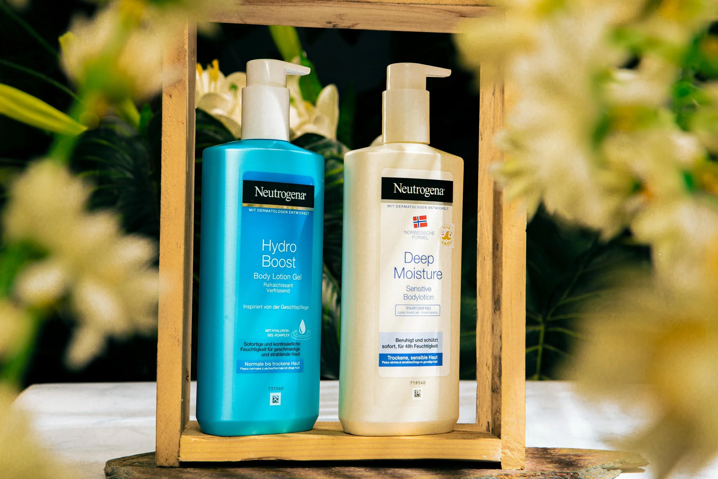 Two bottles of Neutrogena skincare products, including Hydro Boost Body Lotion Gel in blue and Deep Moisture Sensitive Body Lotion in white, placed inside a wooden box with a blurred background of green leaves and white flowers.