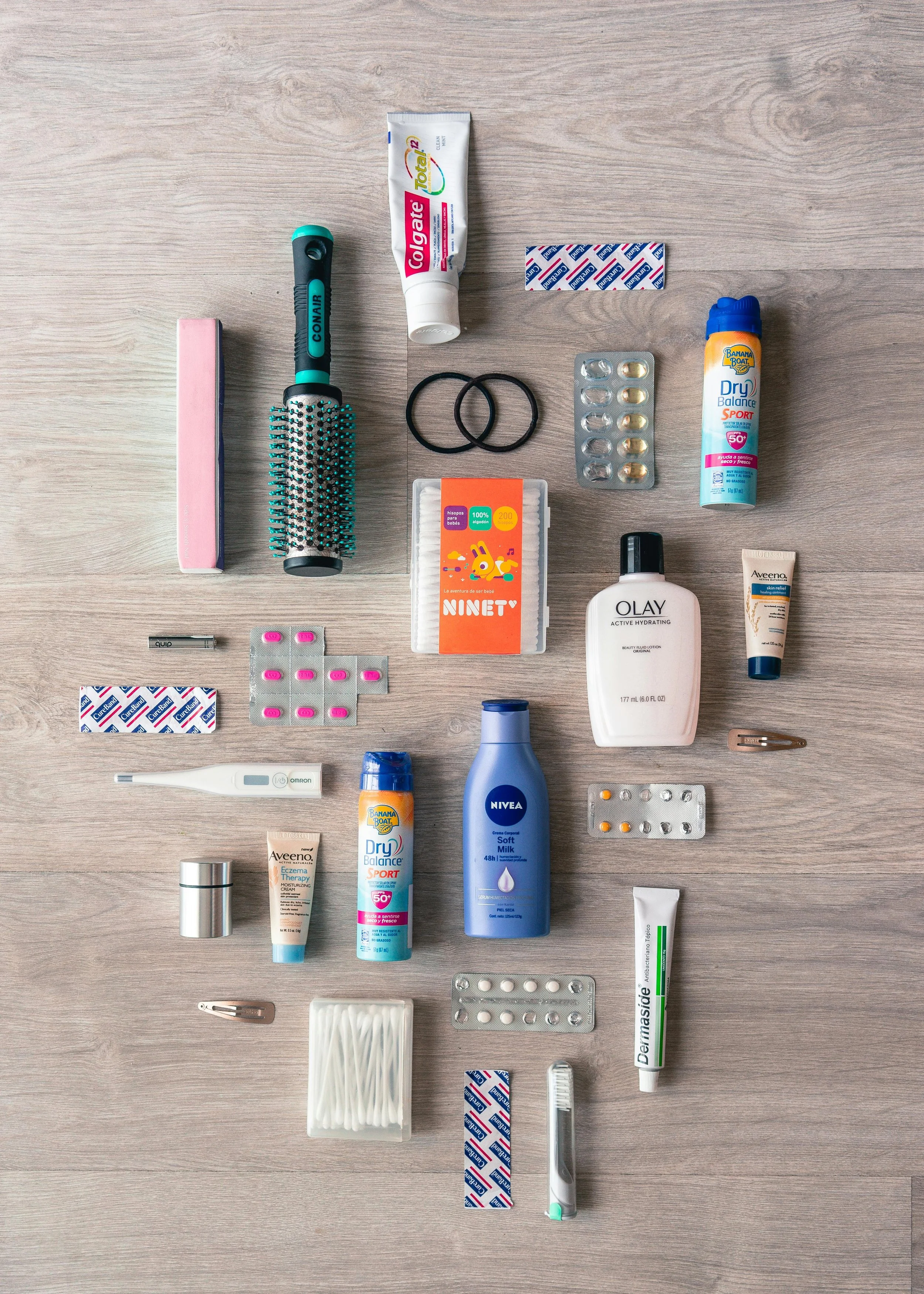 A flat lay of various personal care and health items on a wooden surface, including toothbrushes, hairbrush, toothpaste, medication, sunscreen, moisturizer, and other toiletries.