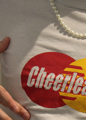 Close-up of a person's chest wearing a Cheerios shirt and a pearl necklace, with part of their hand visible.