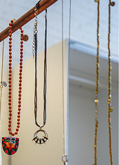 Jewelry hanging on a display rack, including a red beaded necklace, a black necklace with a small charm, and other necklaces with various pendants against a beige background.