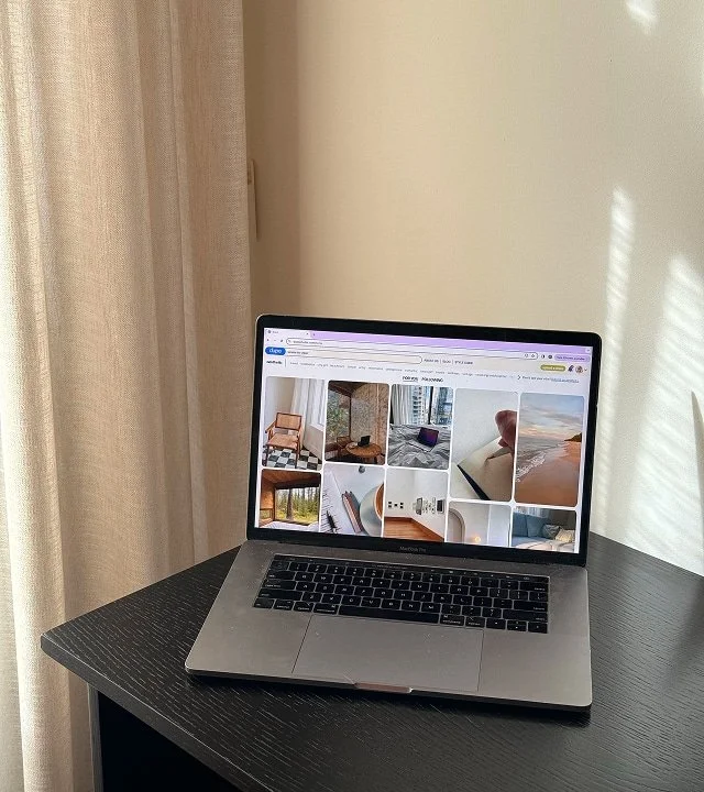 Open laptop on a dark wood table displaying a photo gallery of home interior images, with curtains and a light-colored wall in the background.