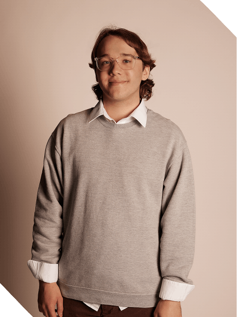 A young man with glasses, wavy brown hair, wearing a light gray sweater over a white collared shirt, standing against a beige background.