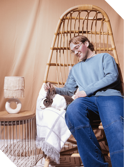 A person with glasses, wearing a light blue sweatshirt and jeans, sitting in a large wicker chair, petting a rabbit.