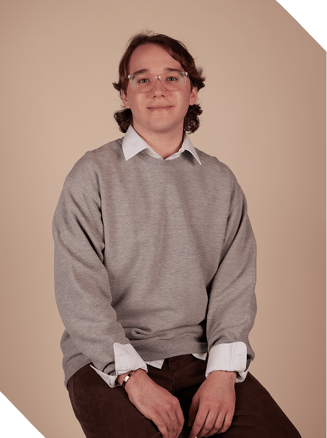 A young man with brown, shoulder-length hair and glasses sitting against a neutral background, wearing a gray sweater over a white collared shirt and brown pants.
