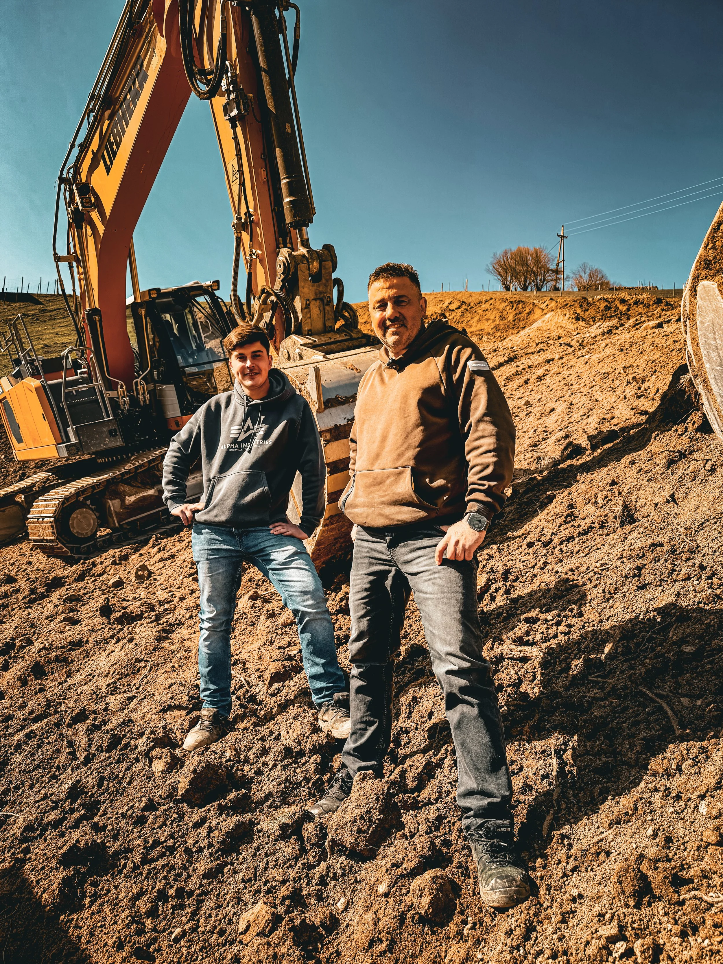 Zwei Männer stehen auf einer Baustelle mit Bagger im Hintergrund, Erde und Steine im Vordergrund, bei sonnigem Wetter.