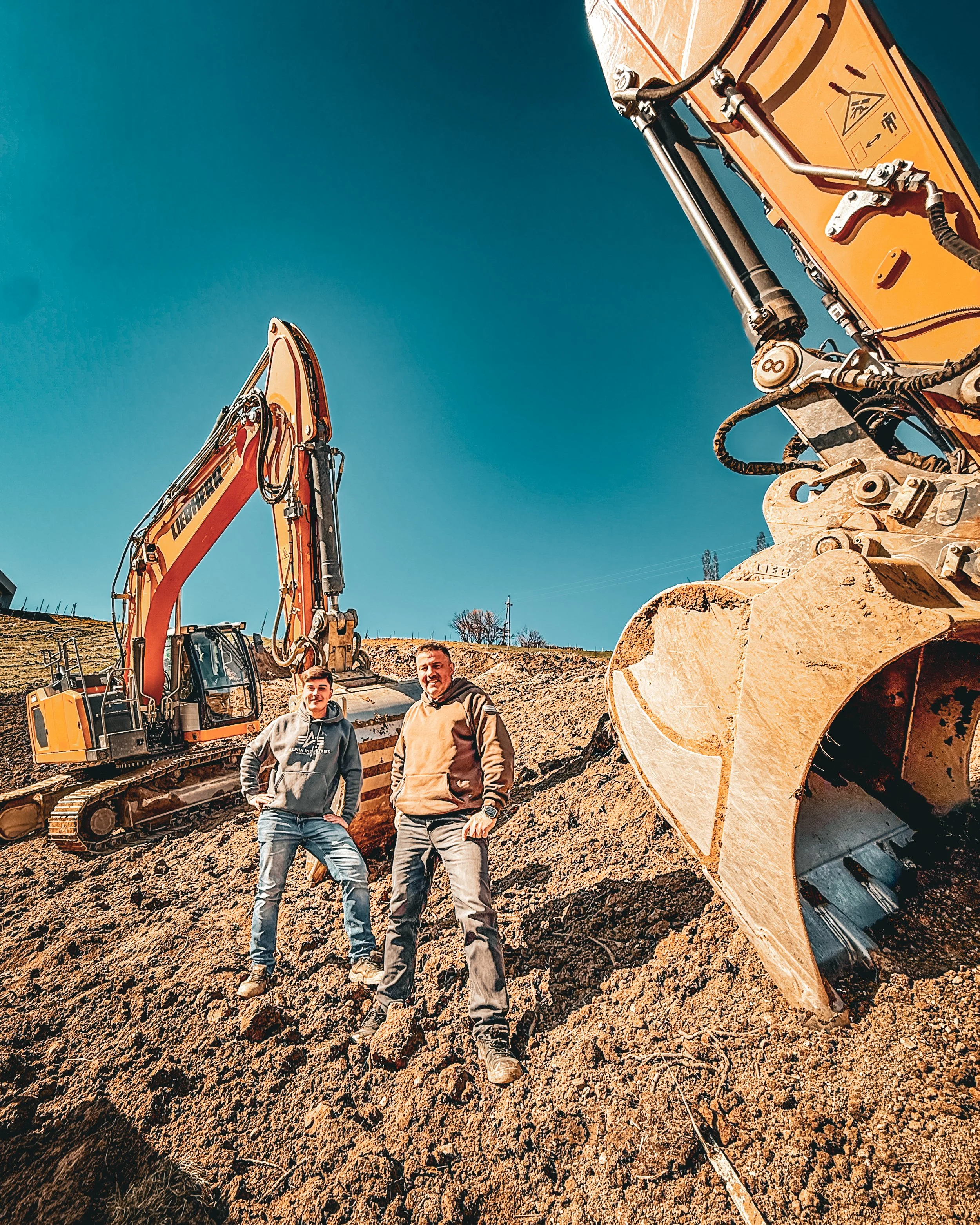 Zwei Männer stehen vor einer Baggermaschine auf einer Erdebaustelle bei sonnigem Himmel.