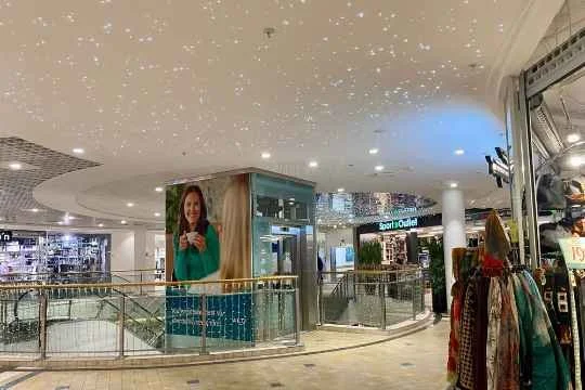 Innendørs utsalg i en kjøpesenter med utsikt mot en reklameplakat som viser en smilende kvinne som holder en kopp. Over er taket dekket med små LED-lys som etterligner stjernehimmel.