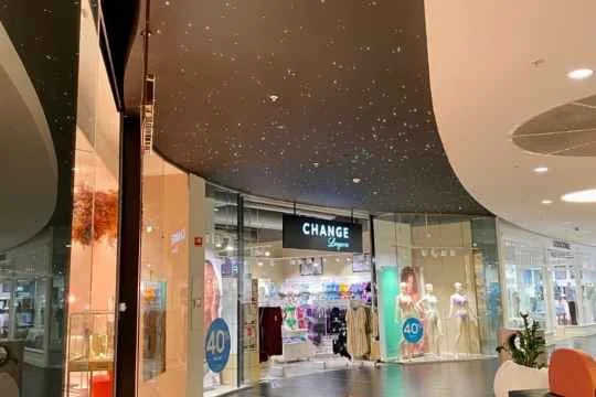 Innvendig utsikt over en klesbutikk i et kjøpesenter, med mannequiner og klær i display, og et digitalt skilt som sier "CHANGE Luxury". Innendørs tak med stjernehimmel effekt.
