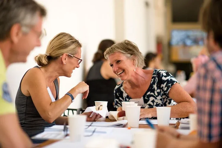 Mehrere Menschen sitzen an einem Tisch in einem Raum, lachen und unterhalten sich. Metaforum Sommercamp