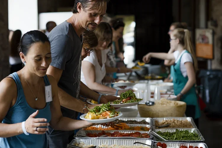 Menschen stehen in einer Reihe beim Buffet und nehmen sich Essen auf Teller, es gibt verschiedene Speisen und Salate.
