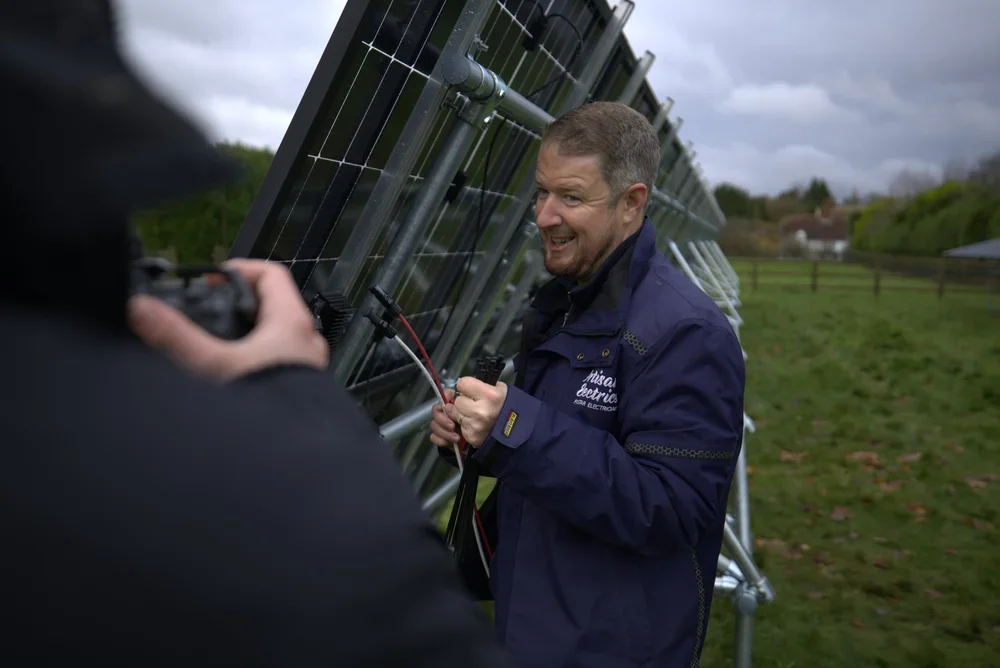 A man in a blue jacket inspecting solar panels outdoors with another person taking a photo