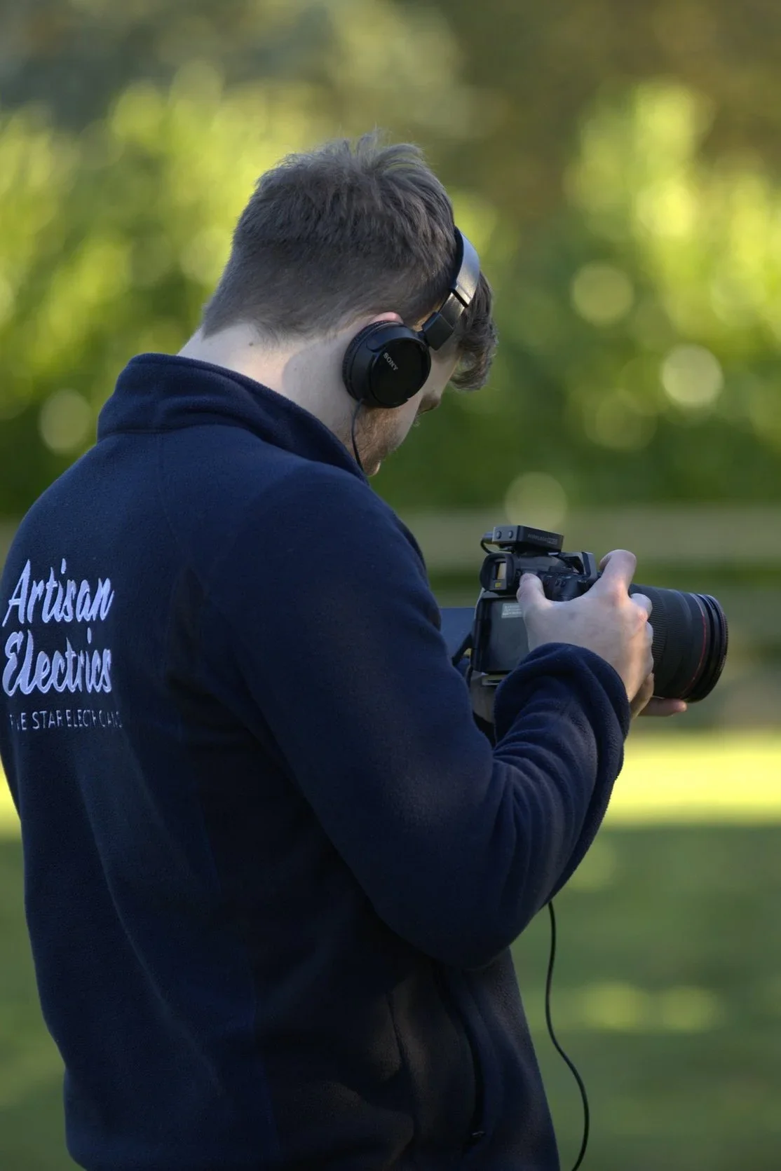 A man wearing a navy blue hoodie with 'Artisan Electric' written on the back is holding a camera and looking at its screen, outdoors with blurred green trees in the background.
