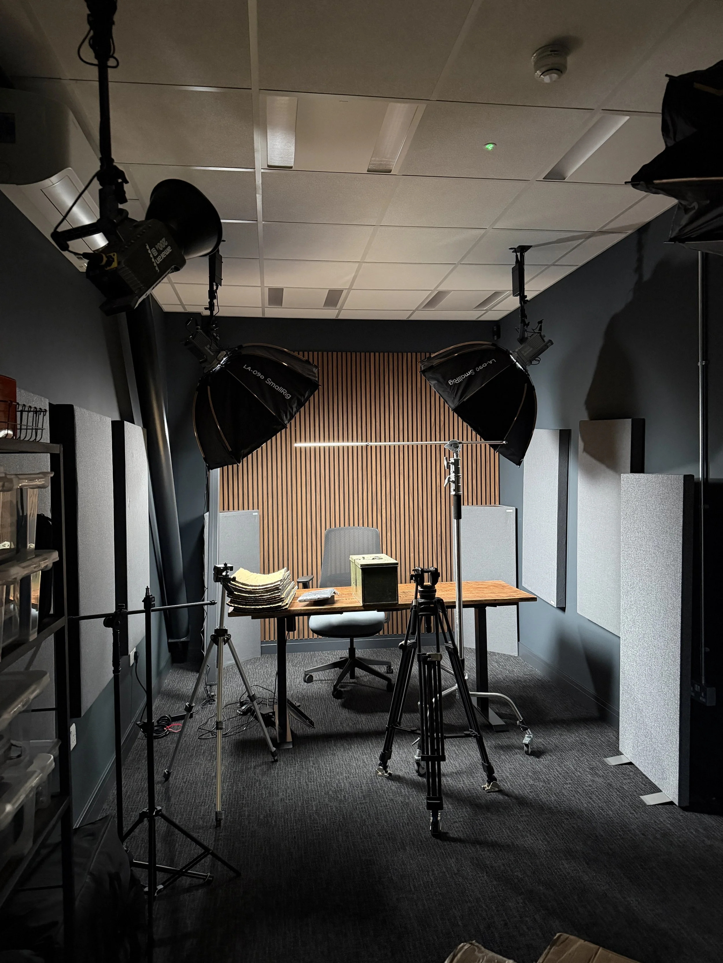 A photography studio with professional lighting and equipment facing a desk with a chair, papers, and a small box, set up for a recording or interview.