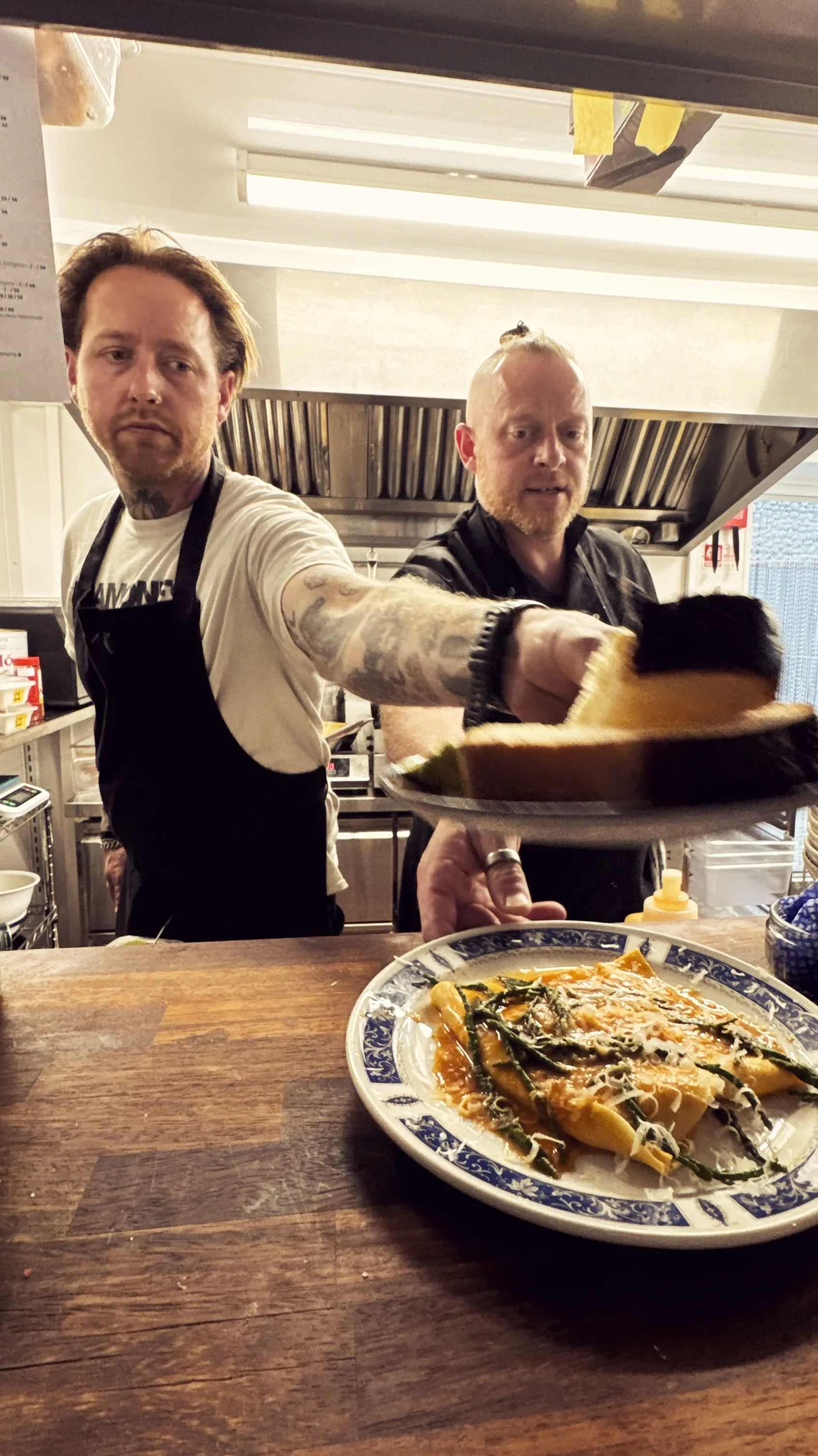 Two male chefs in a restaurant kitchen preparing a dish of lasagna with vegetables, one handing the plate to the other, with a pizza in the foreground.