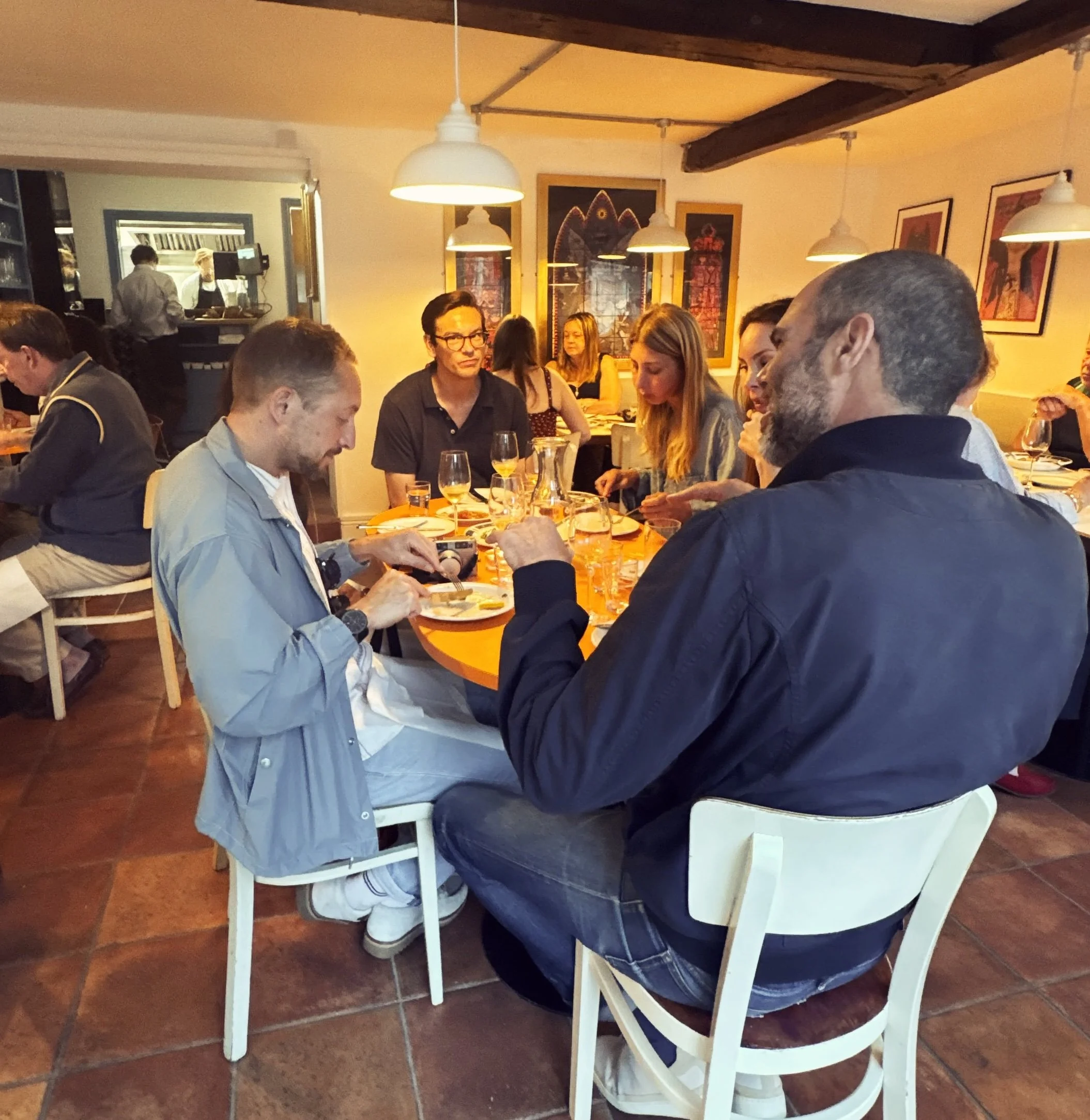 People sitting around a dining table in a restaurant, with some engaging on their phones and conversations, decorated with framed artwork on the walls and hanging pendant lights.