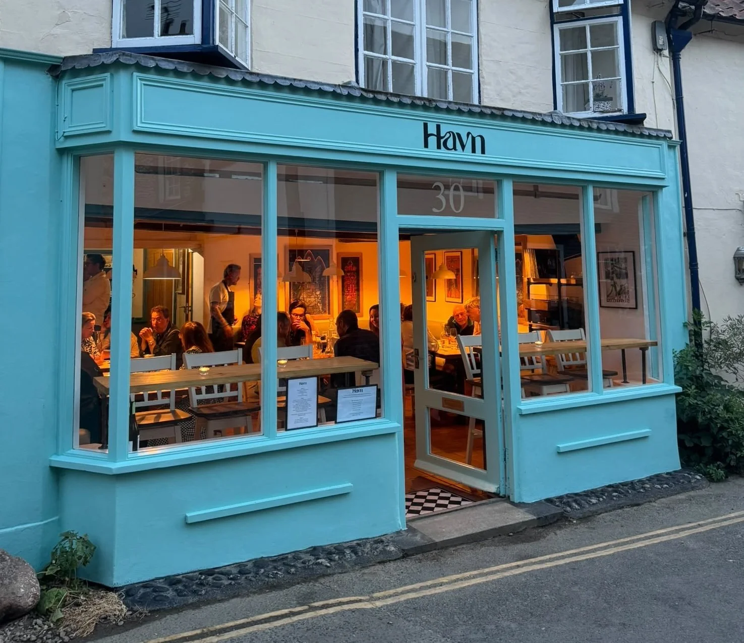 A colorful cafe storefront with large windows, showing people dining inside. The exterior is painted bright turquoise, with the name 'Havn' displayed above the entrance. Inside, warm lighting illuminates the patrons and artwork on the walls.