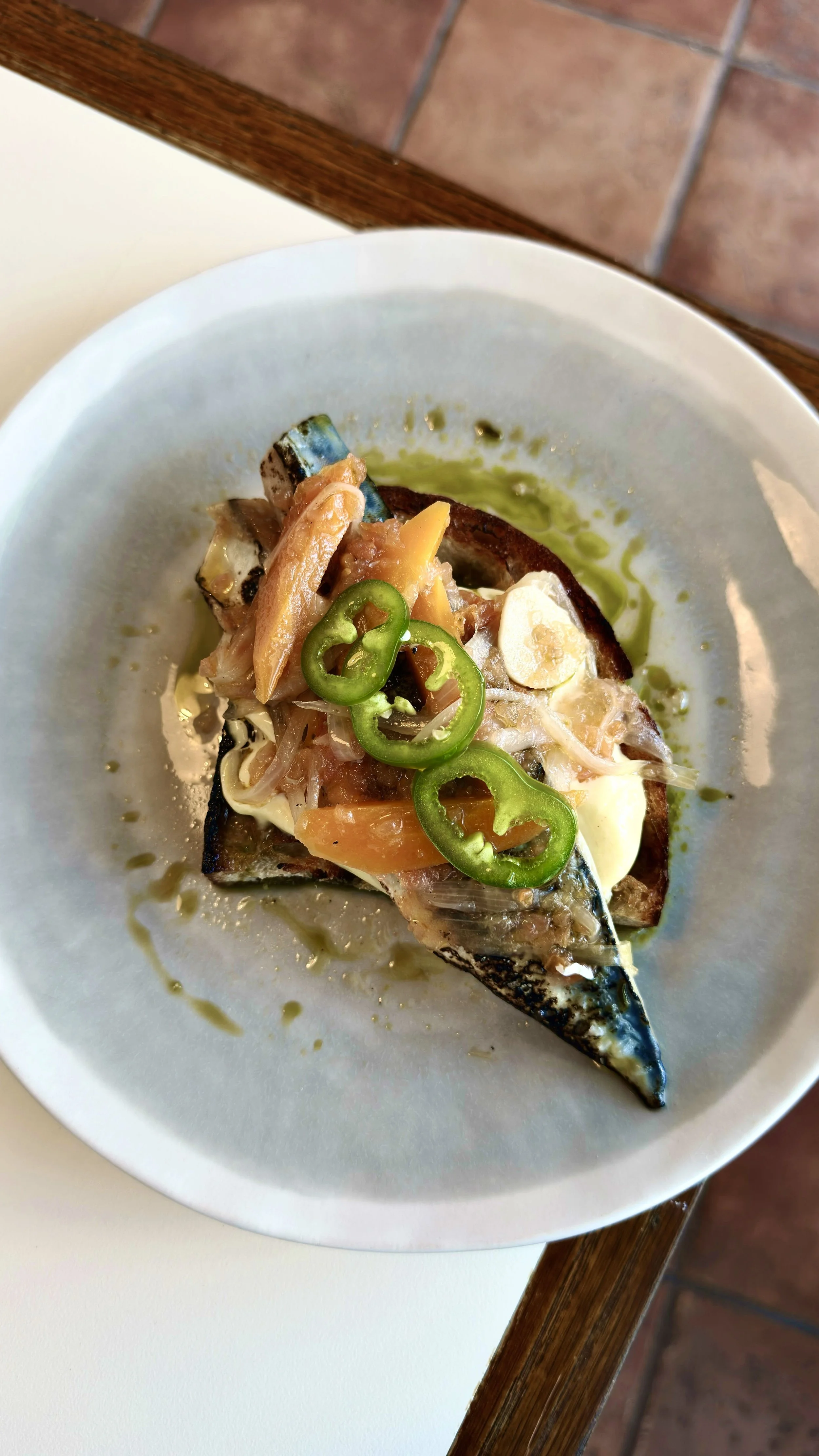 A dish of grilled fish topped with sliced jalapenos, tomatoes, onions, and herbs, drizzled with green oil.