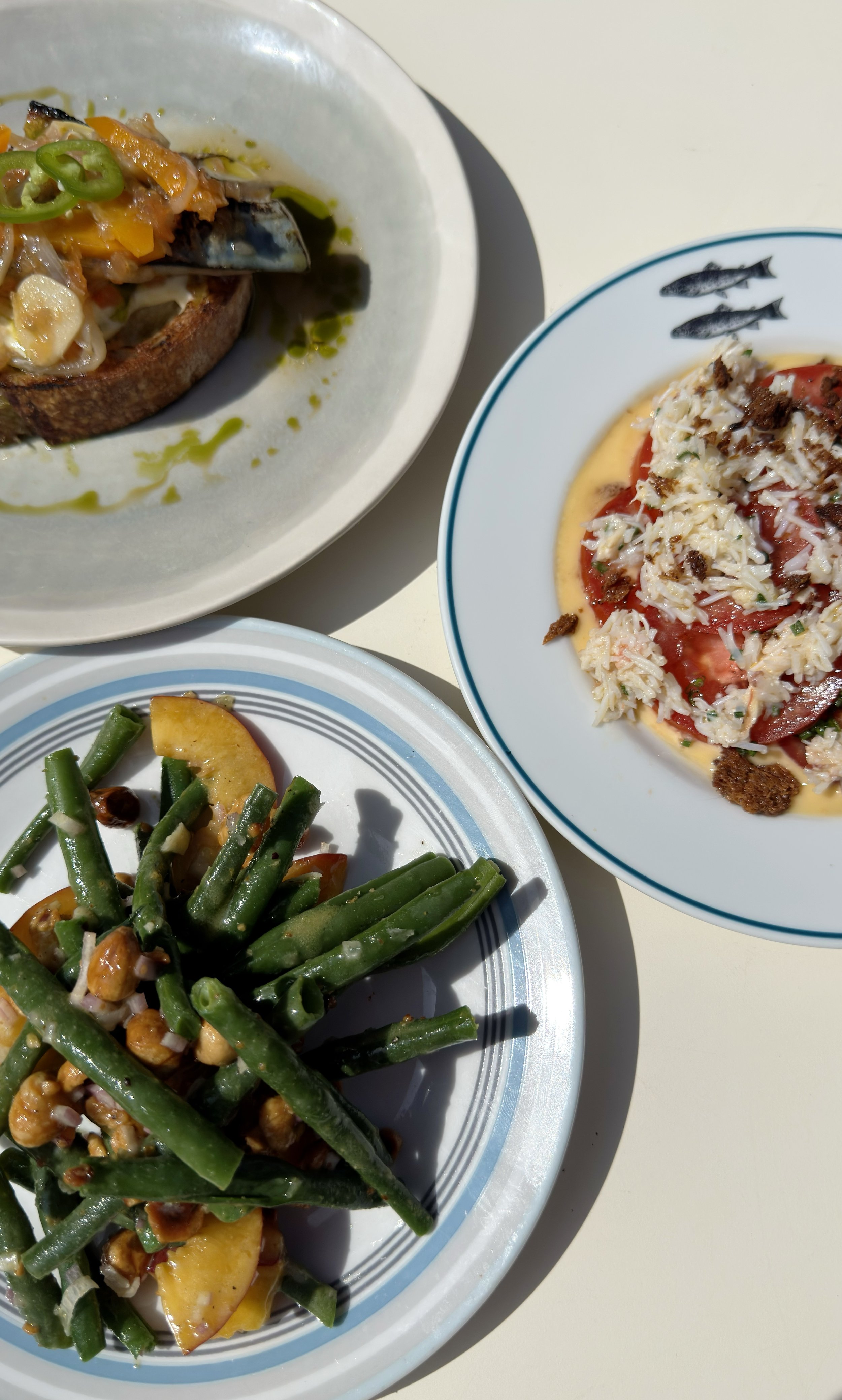 Three plates of food: one with green beans and yellow potatoes, one with a crab dish topped with sauce and cheese, and one with an open-faced sandwich with tuna, tomato, cheese, and sauce.