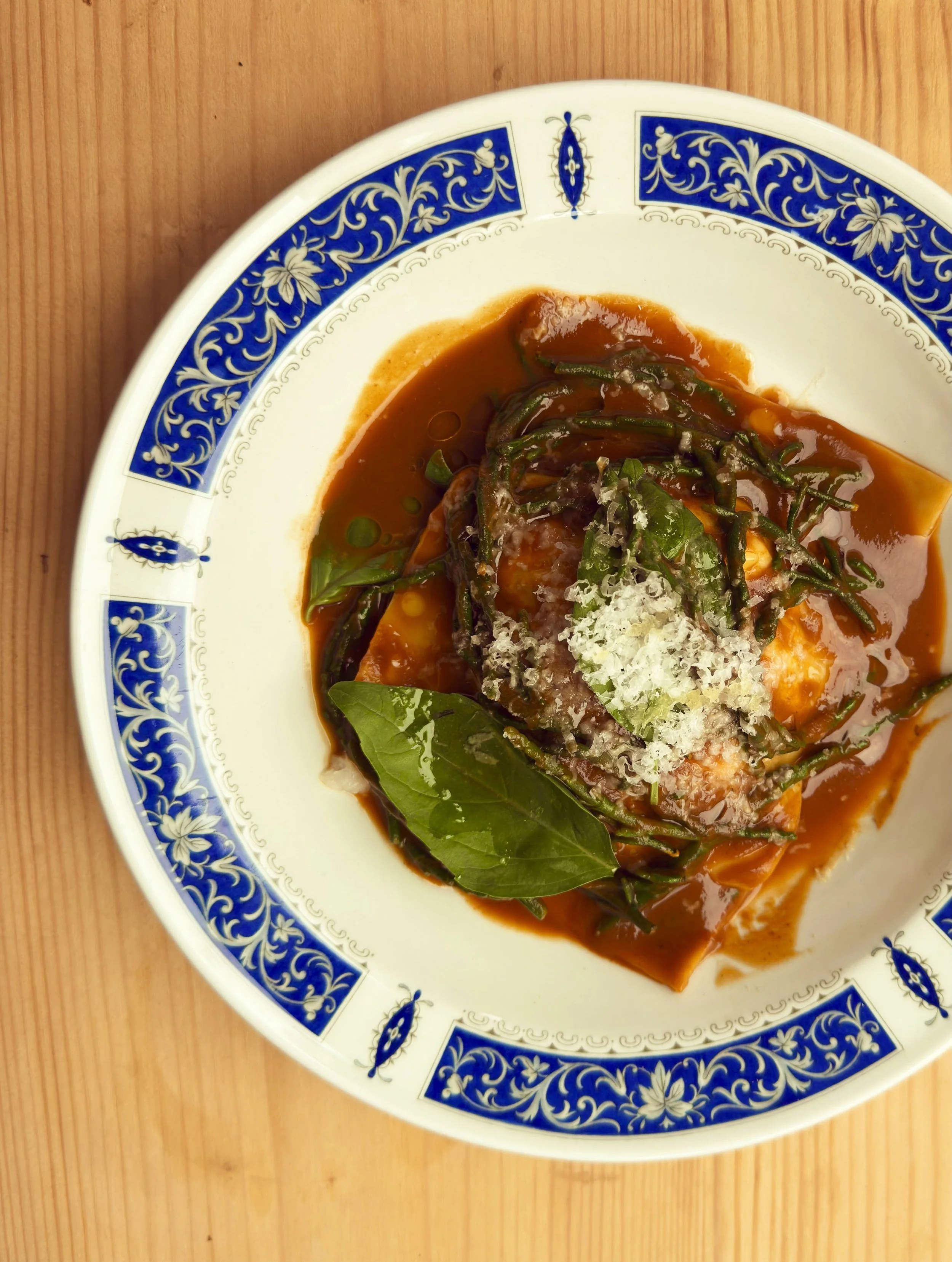A plate of Italian lasagna with tomato sauce, topped with grated cheese, basil leaves, and green beans, on a blue and white ornate ceramic plate placed on a wooden surface.
