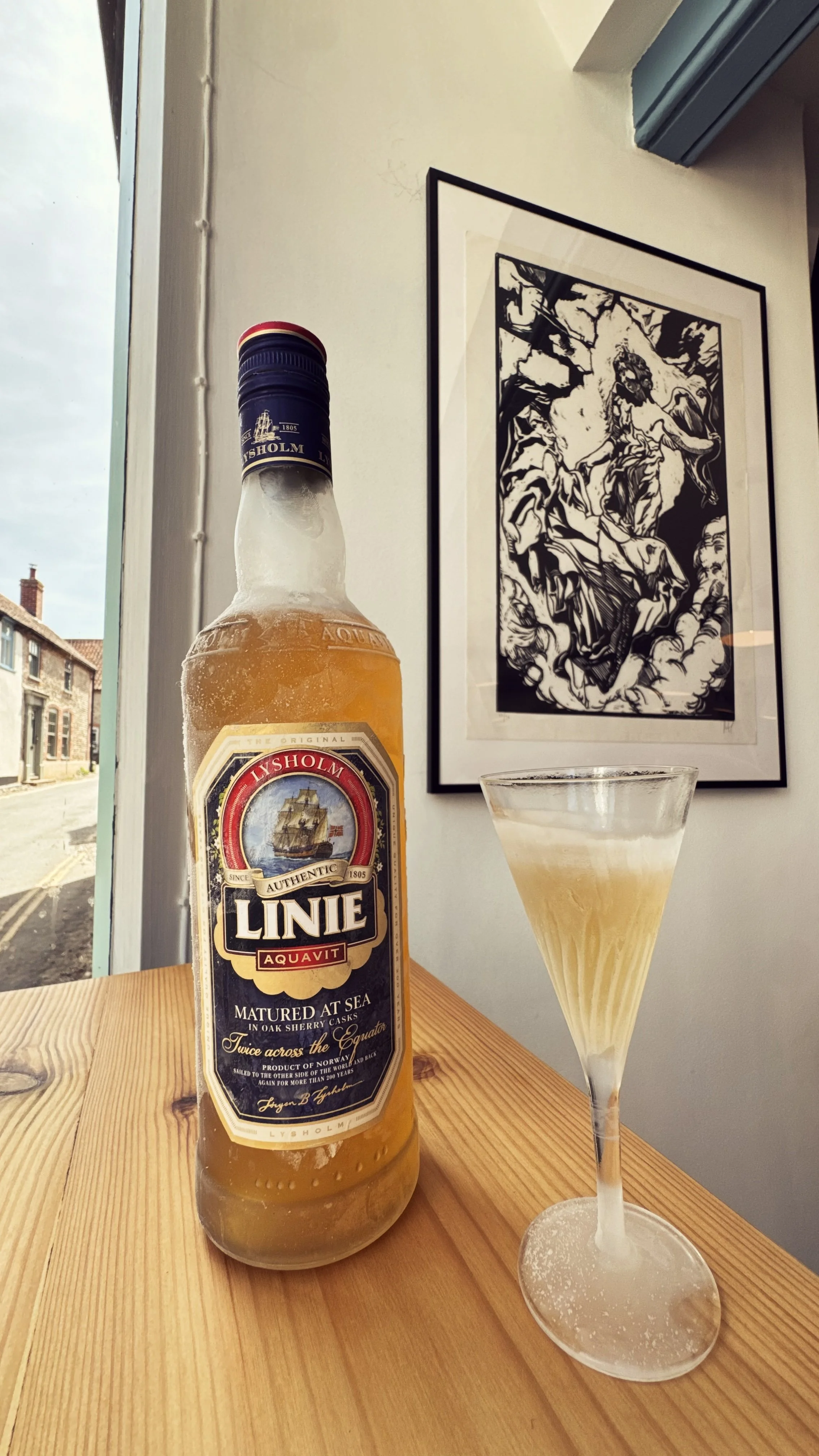 A glass of pale, frothy beer and a bottle of Lysholm Linie Aquavit on a wooden table near a window with a black and white art print on the wall in the background.