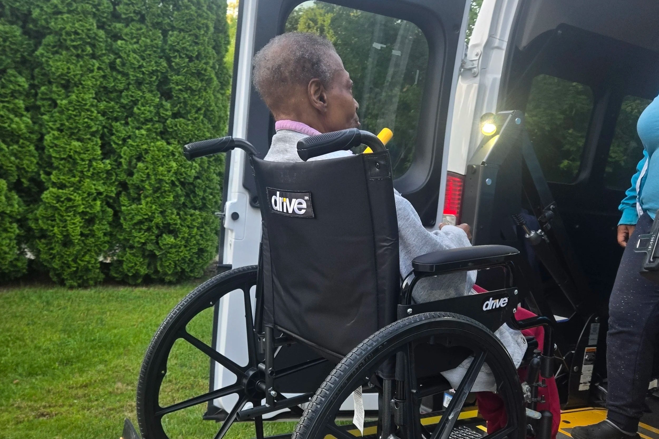 An elderly man sitting in a wheelchair with a black backrest. He was gently support to enter the vehicle.