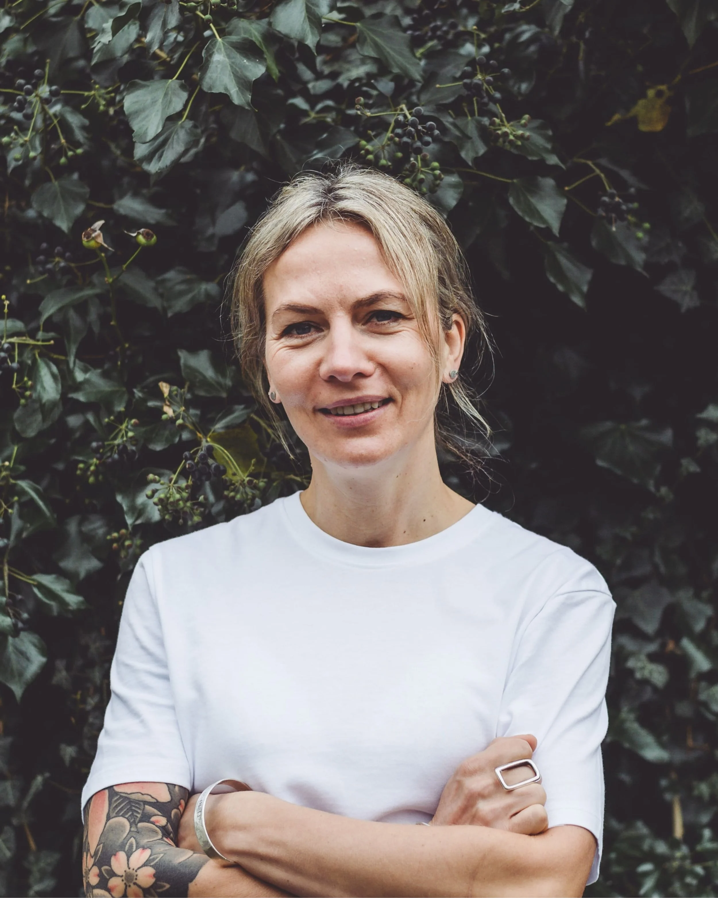 A woman with blonde hair and tattoos on her arm, wearing a white t-shirt, standing in front of green leafy bushes.
