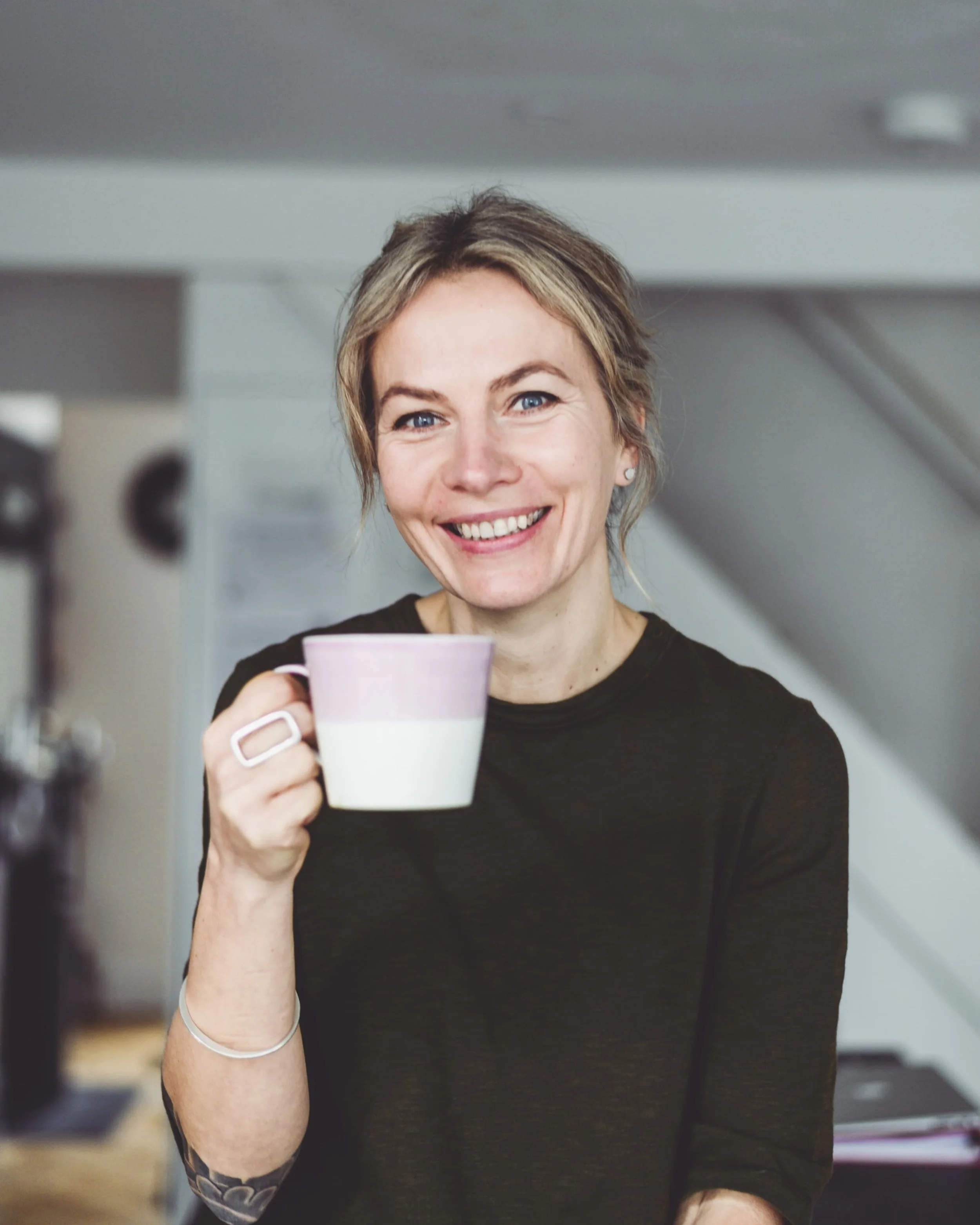 A woman with short blond hair and tattoos on her left arm, smiling and holding a mug, standing in an indoor setting.
