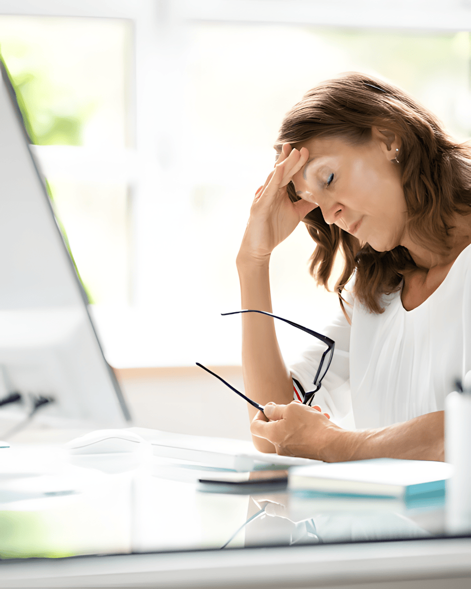 A woman sitting at a desk holding her head in frustration, with glasses in her other hand, in an office setting with natural light.
