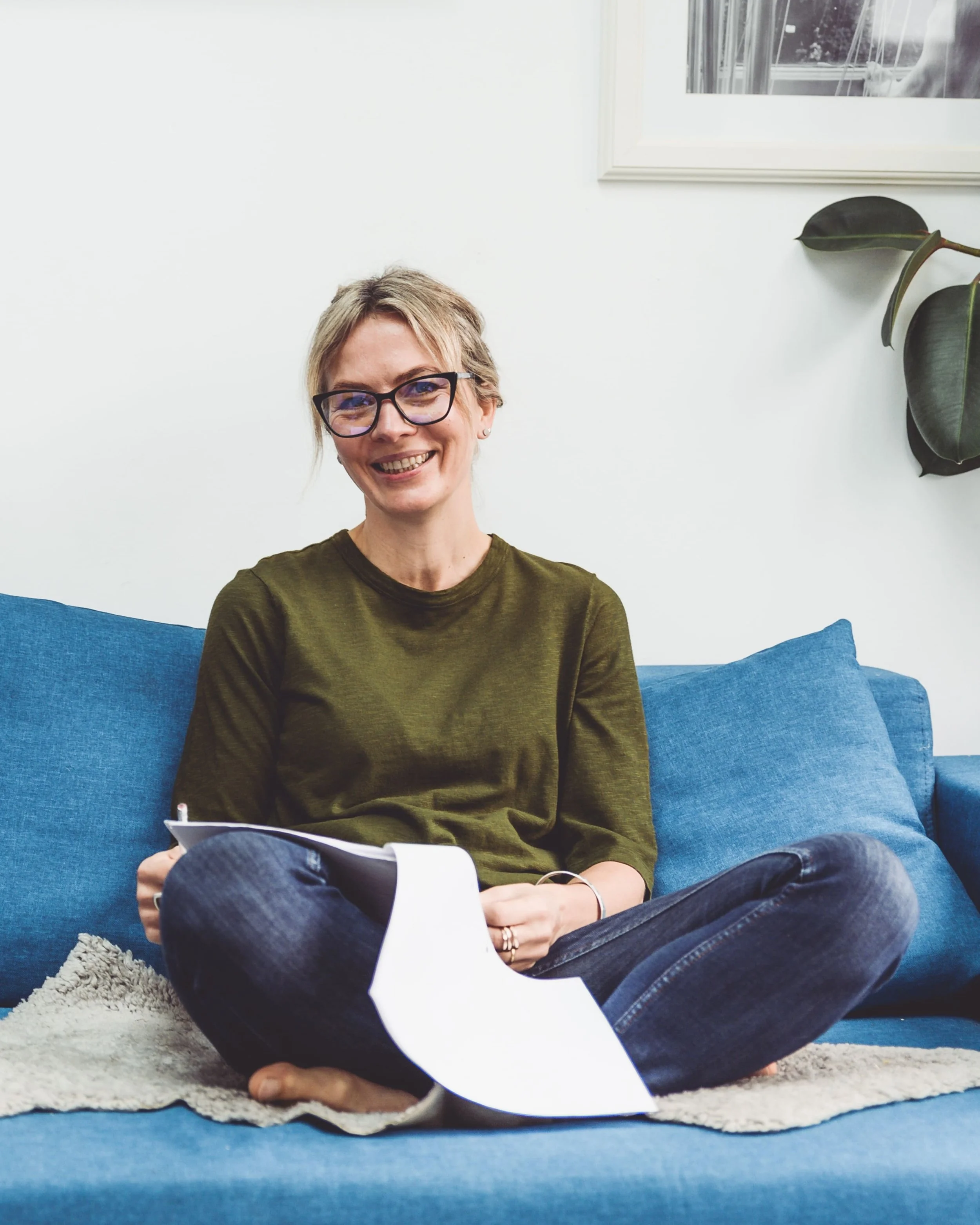 Woman with short blonde hair and glasses smiling, sitting cross-legged on a blue couch, holding papers in her hands, with a plant and framed picture in the background.