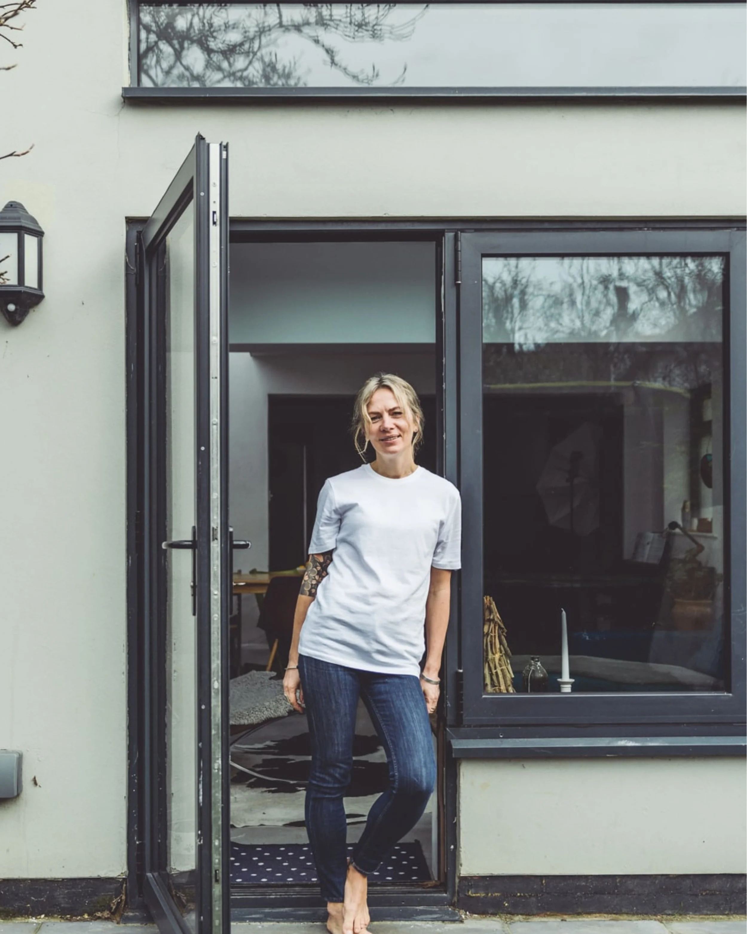 A woman standing barefoot at a door threshold, smiling, with a tattoo on her left arm, wearing a white T-shirt and dark jeans, in front of a modern house with large windows.