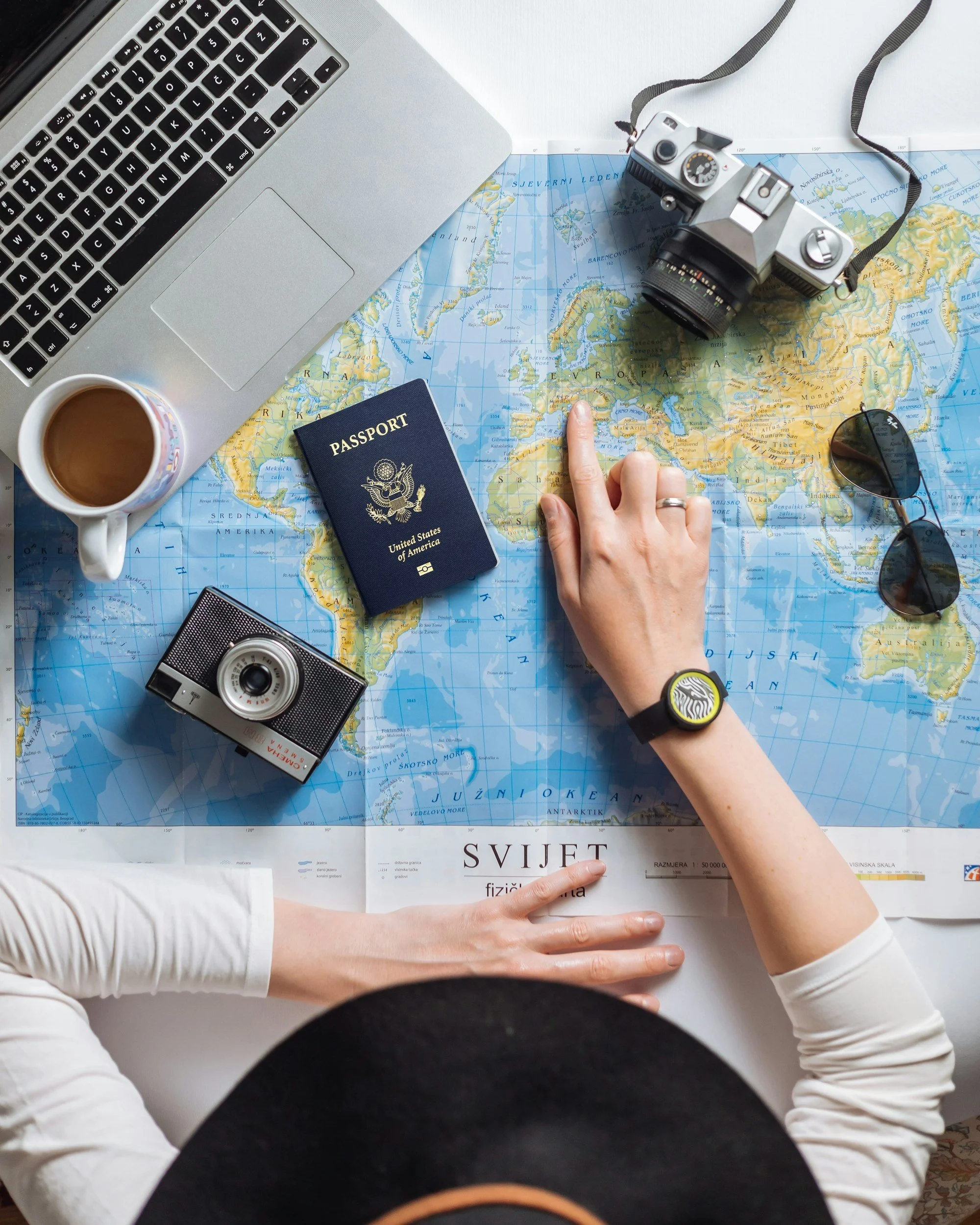 Overhead view of a person wearing a white long-sleeve shirt and black vest, pointing at a world map with one finger. On the map, there are a silver vintage camera, a pair of black sunglasses, a blue United States passport, a white coffee cup filled with coffee, and two other cameras. A silver laptop is also visible on the left side of the image.