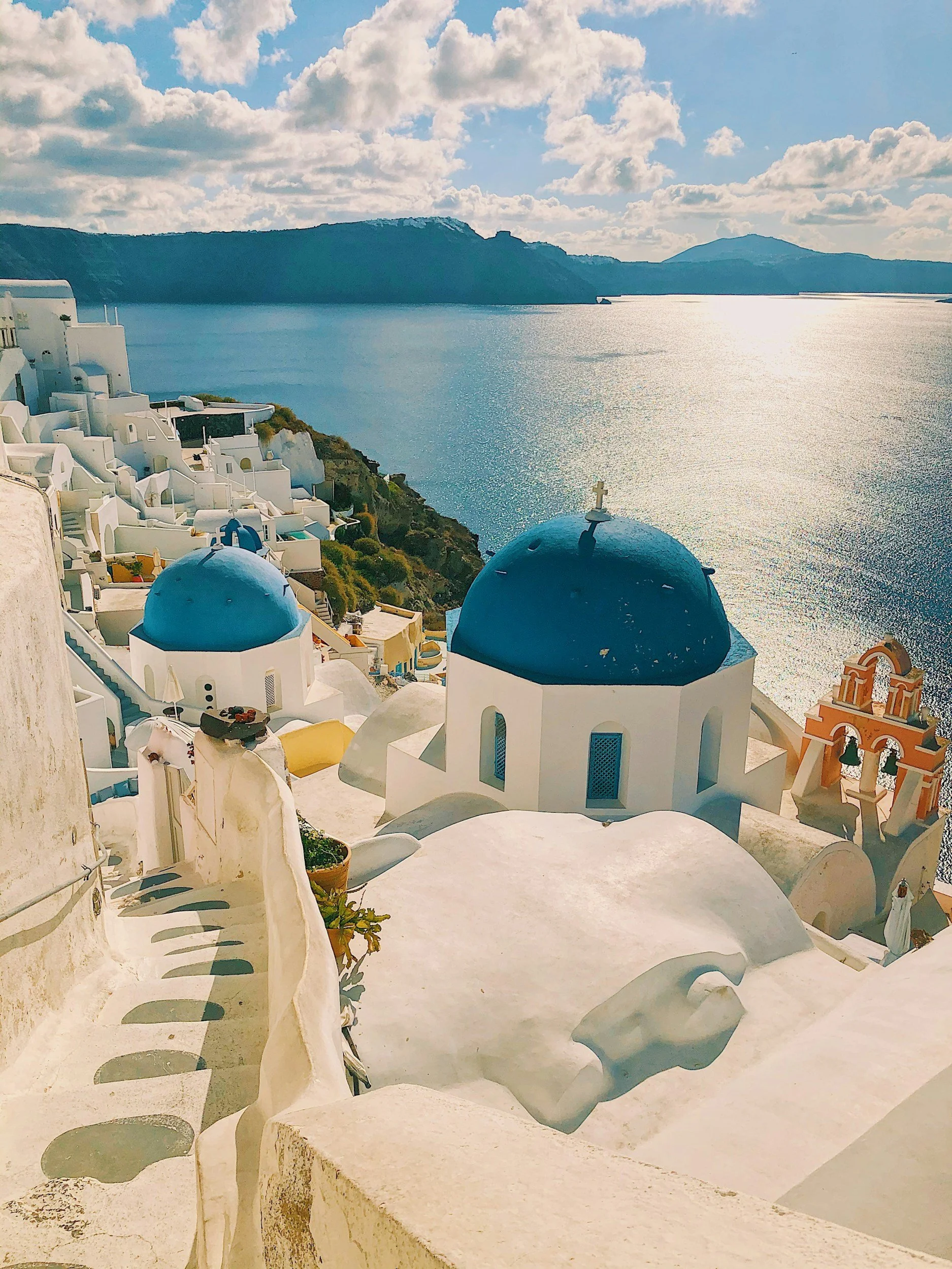 View of Santorini, Greece showing white buildings with blue domes, the Aegean Sea in the background, and a partly cloudy sky.