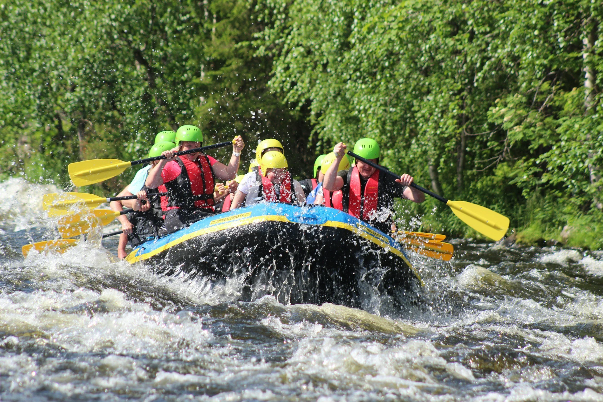 Group of people white-water rafting on a river with green trees in the background, wearing helmets and life jackets, paddling in a blue raft.