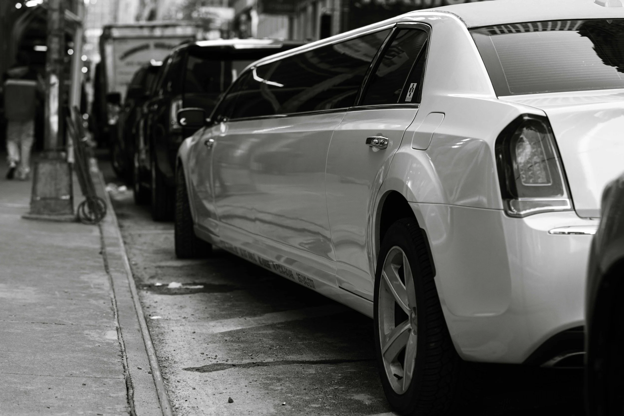 A row of parked cars on the street in an urban area, with a white car in the foreground among darker colored vehicles.
