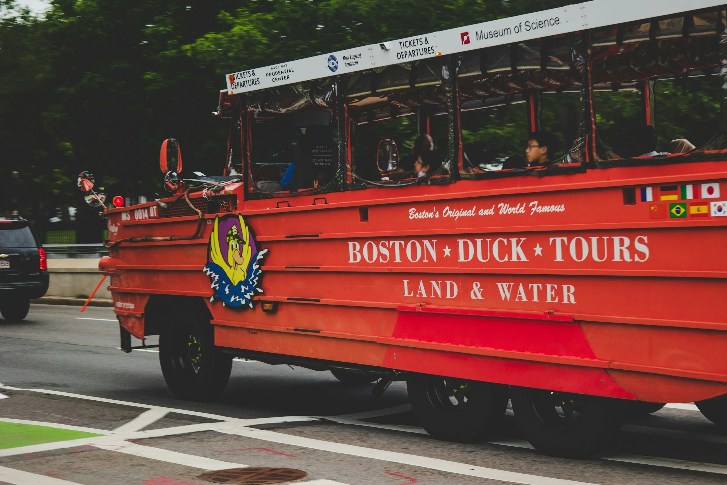 Red amphibious tour bus for Boston Duck Tours driving on city street with trees in background.