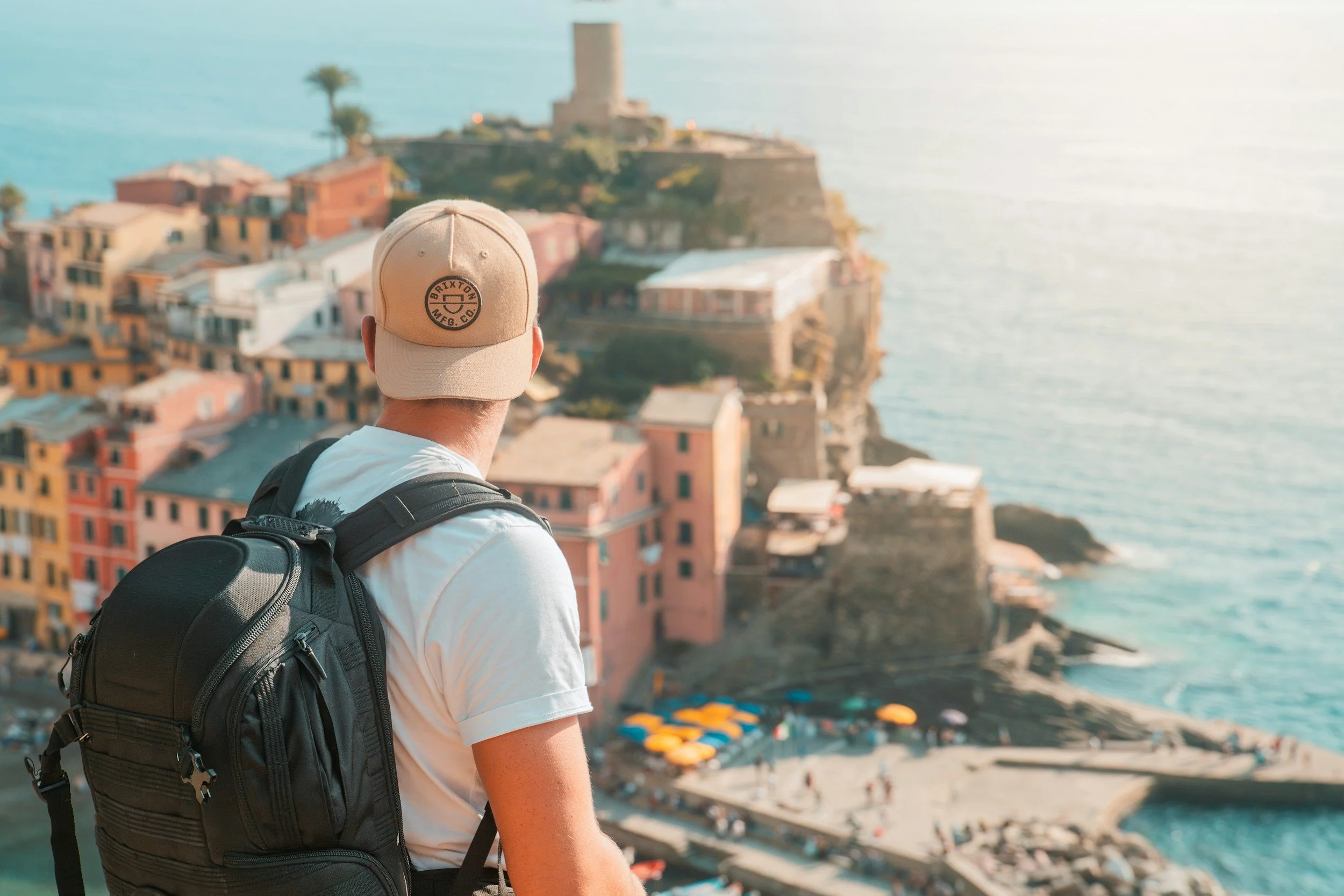 A man wearing a beige cap with the words 'BRIXTON MFG. CO.' and a circular logo, a white t-shirt, and a black backpack, looking at pastel-colored buildings and a coastal landscape with a tower structure, ocean, and beach umbrellas in the background.