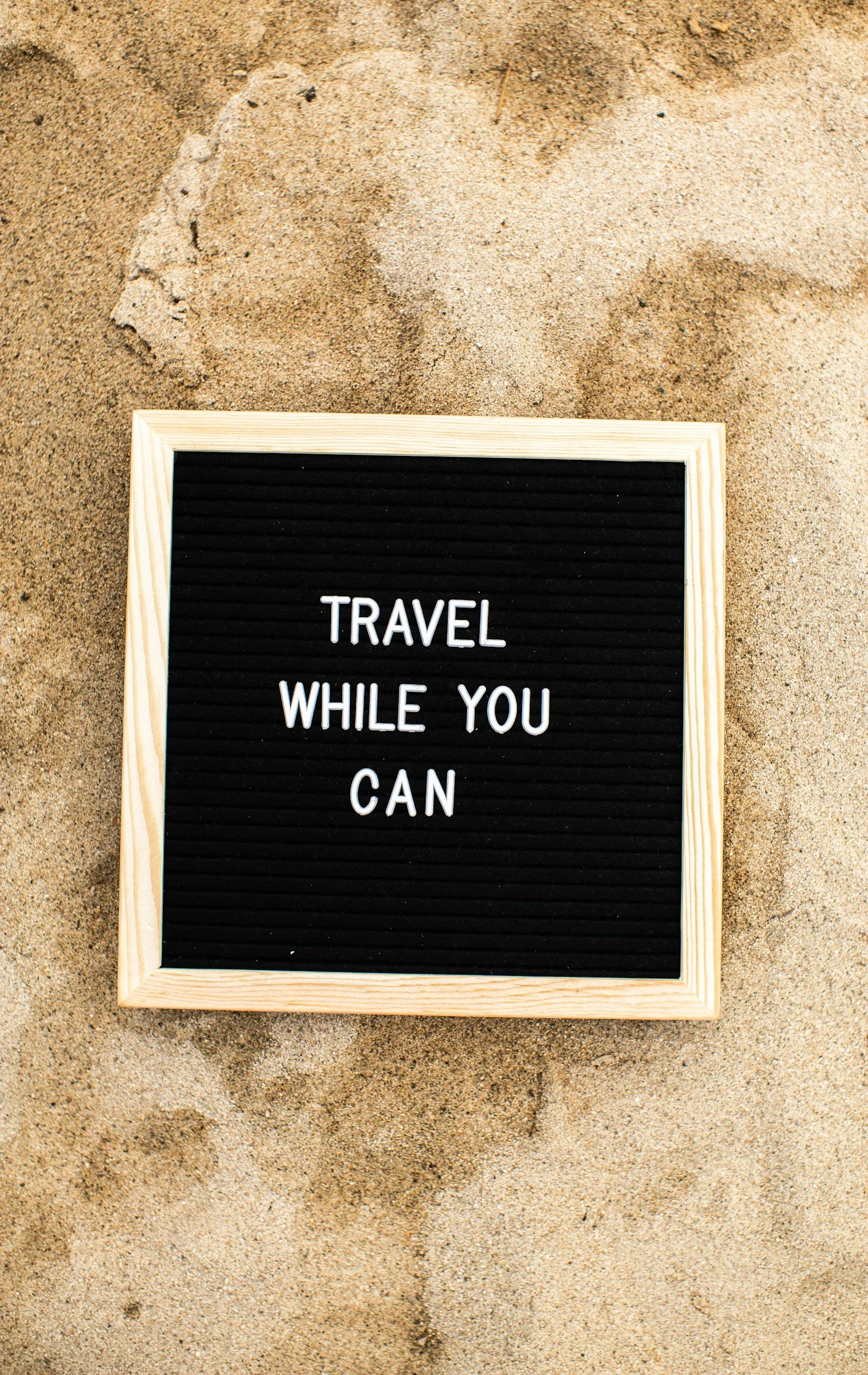 A black letter board with white text saying "Travel While You Can" is placed on sandy ground.