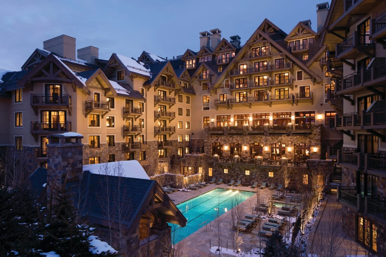A large lodge-style hotel with multiple stories, warm lighting, and balconies overlooking a swimming pool. Snow is present on the rooftops and surrounding trees at dusk.