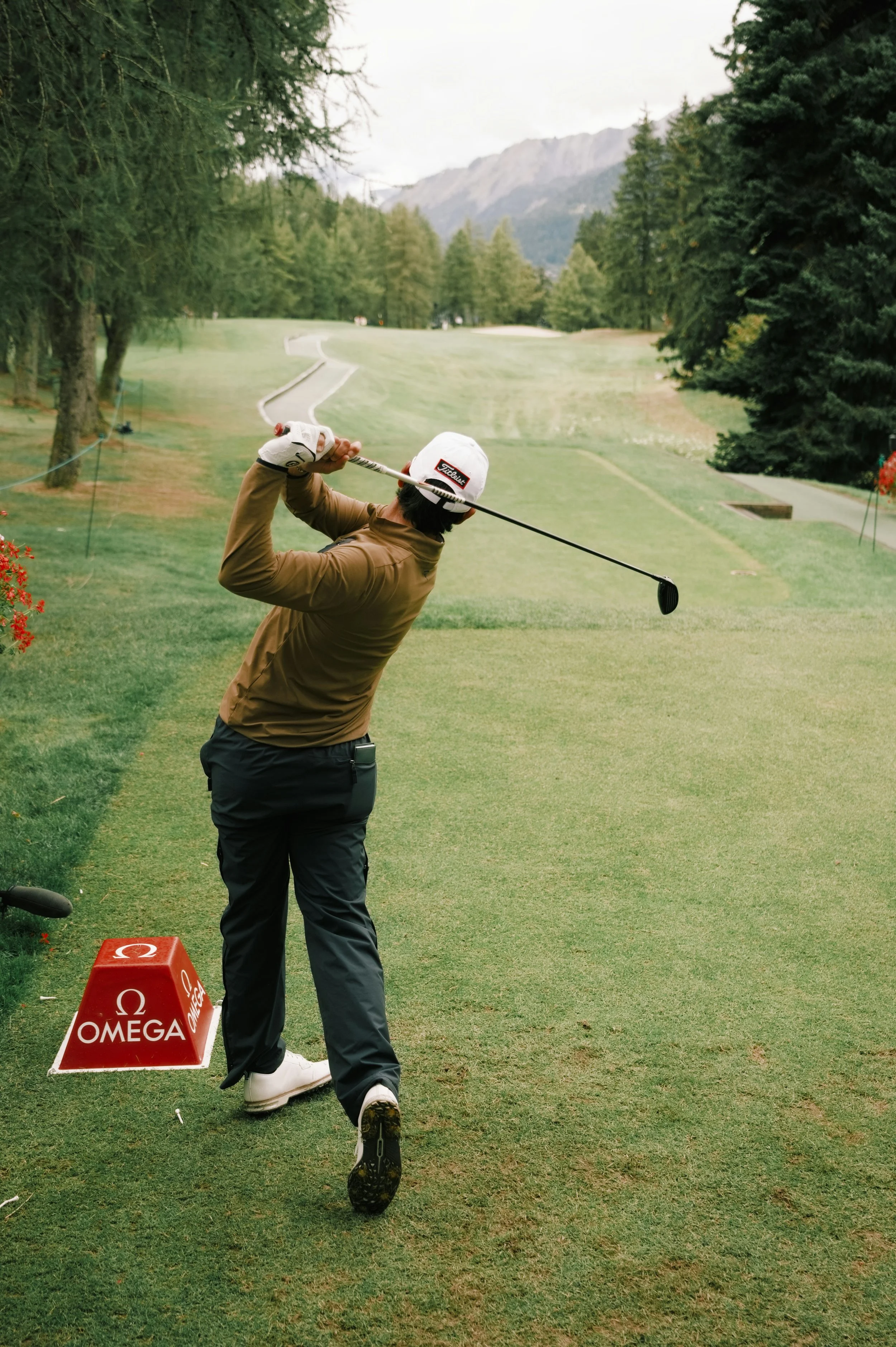 A golfer in a brown jacket and cap swings a golf club on a golf course. There are trees and mountains in the background, and a red Omega branded sign on the ground nearby.
