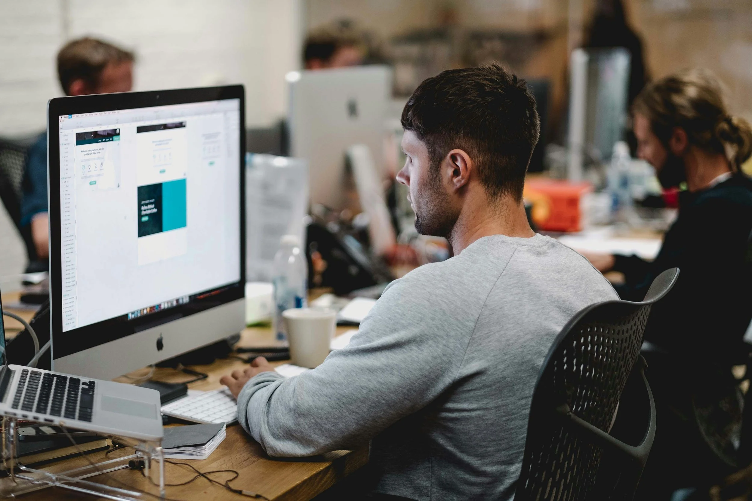 Man working at a computer in an office with other people working in the background.