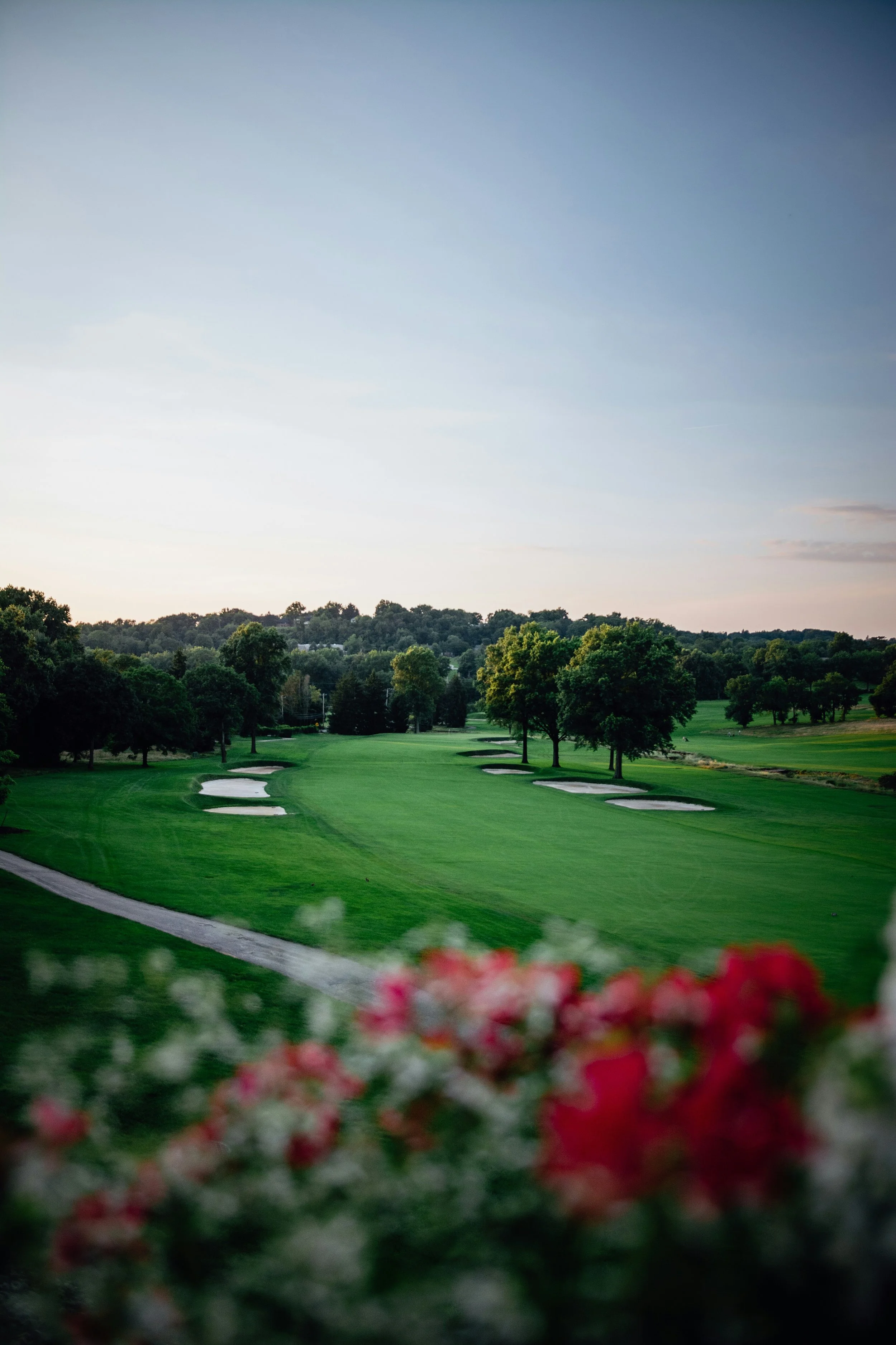 A scenic view of a golf course with green grass, sand bunkers, trees, and a distant hill under a clear sky.