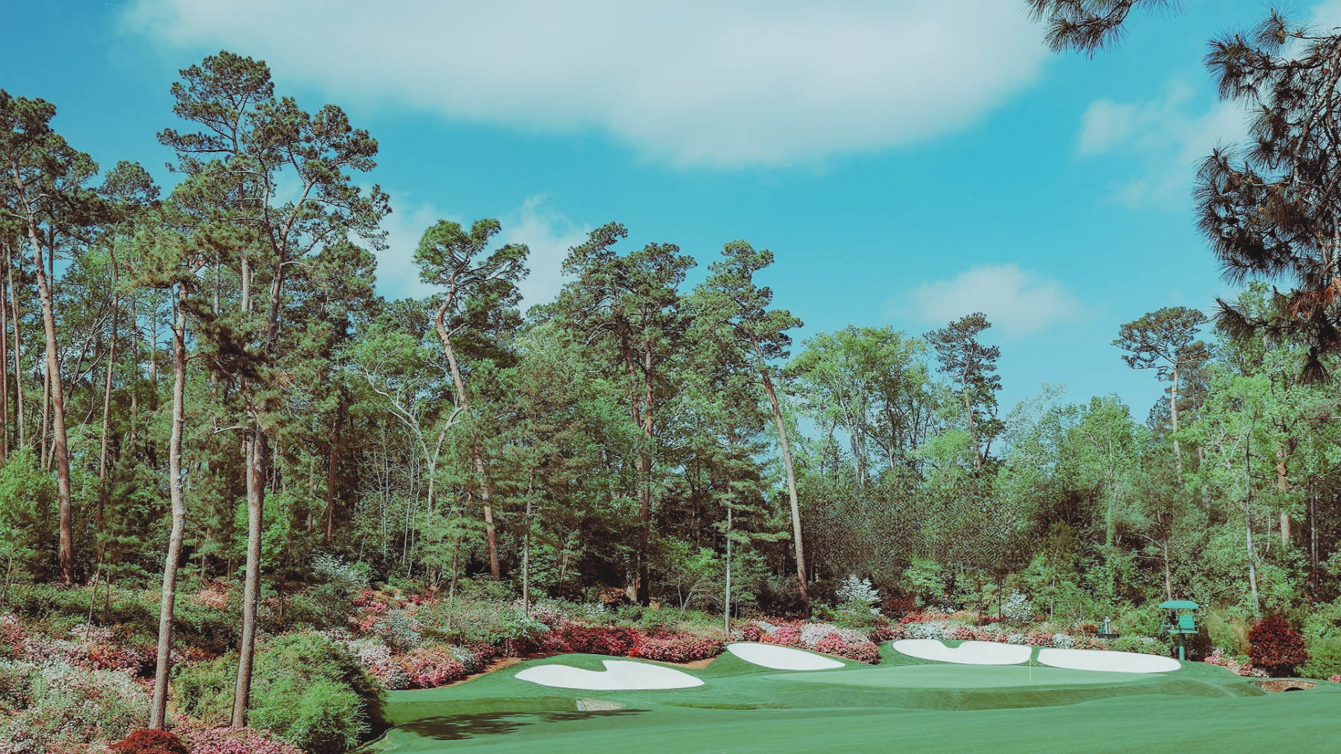 Golf course with sand bunkers and a flag, surrounded by tall trees and colorful bushes under a partly cloudy sky.