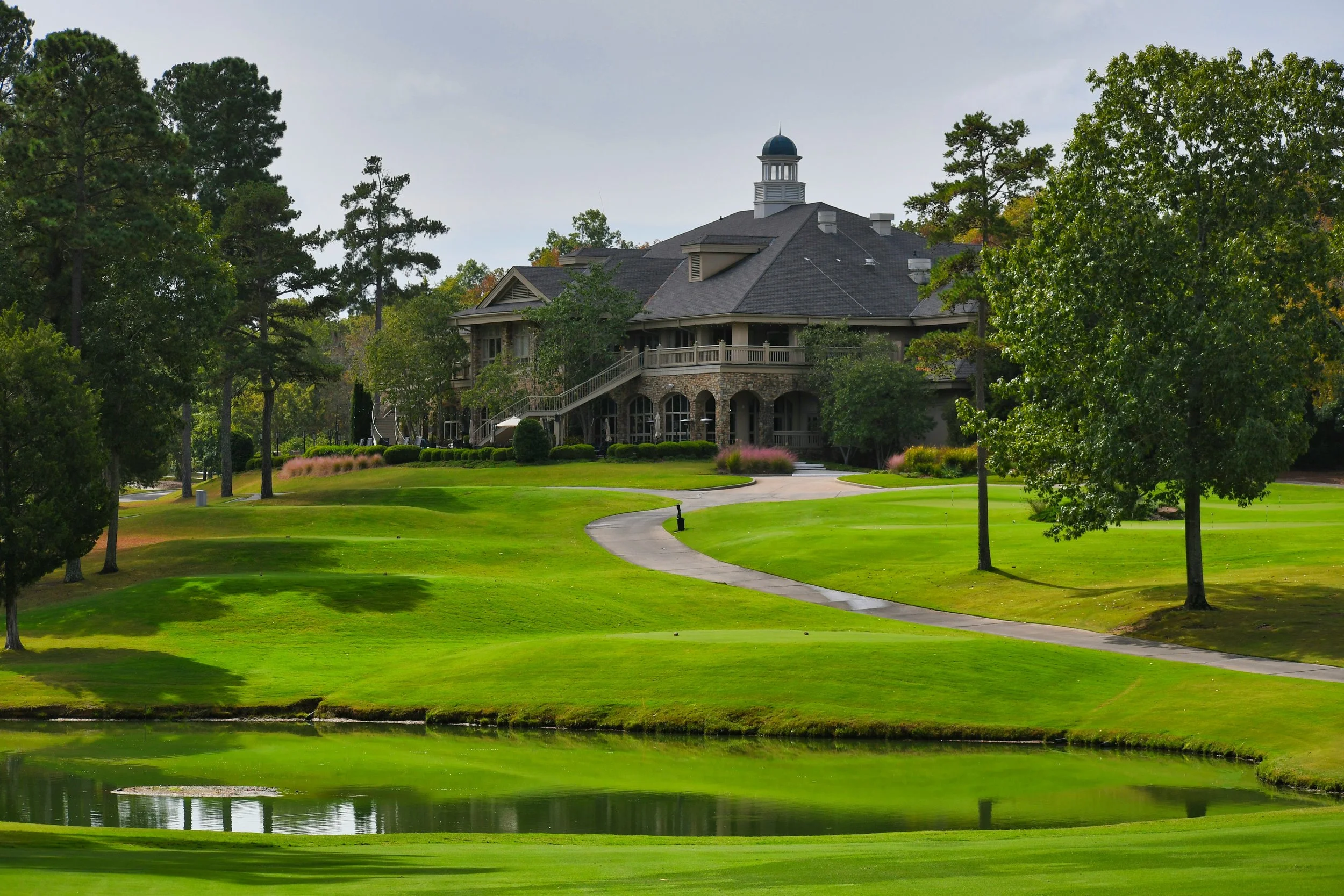 A large house surrounded by tall trees on a well-maintained golf course with green grass, winding paved paths, and a small pond in the foreground.