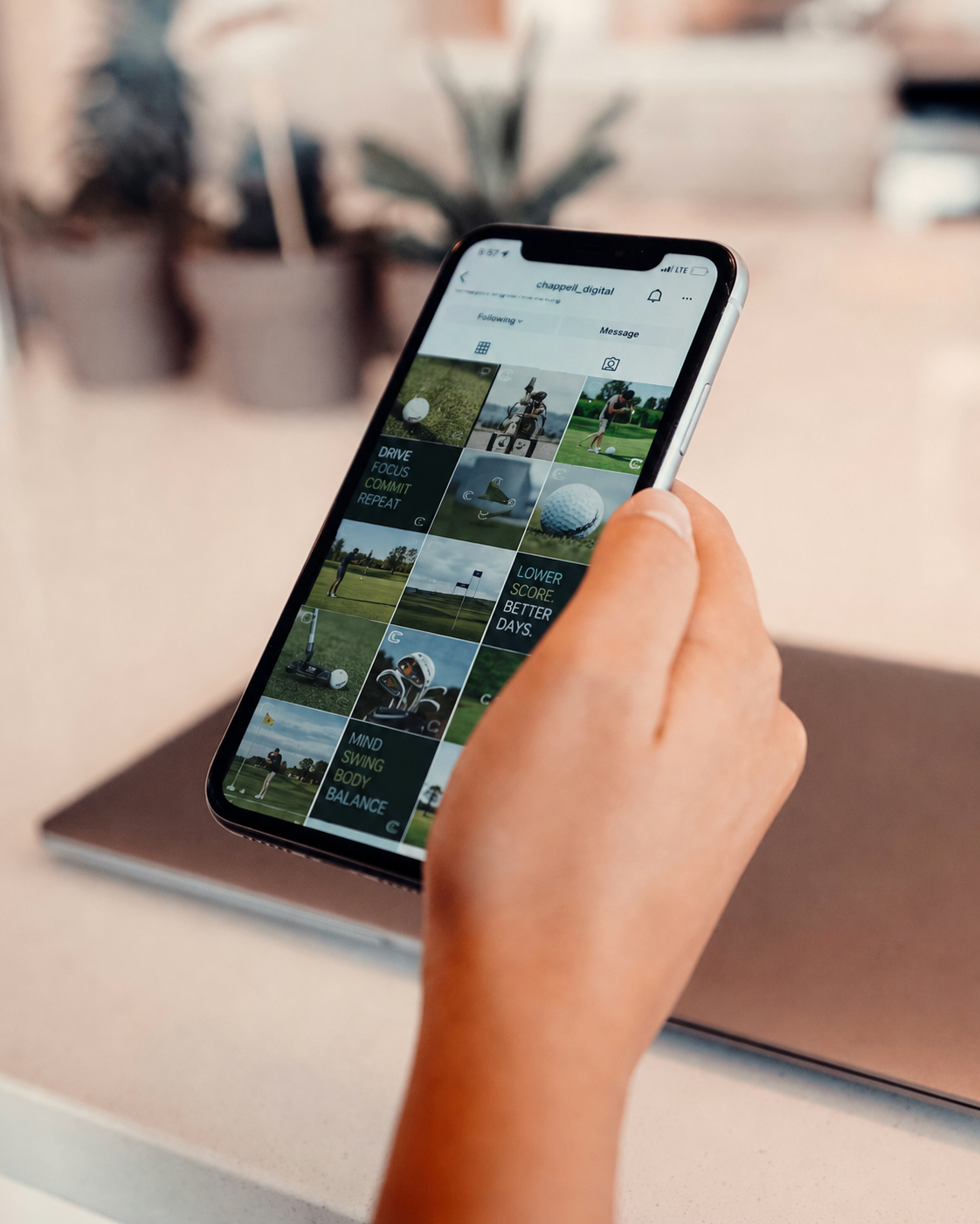 A person holding a smartphone displaying an Instagram profile page with various social media-related posts. The person's arm has multiple bracelets, and they are sitting on a wooden surface with a patterned cloth or blanket nearby.