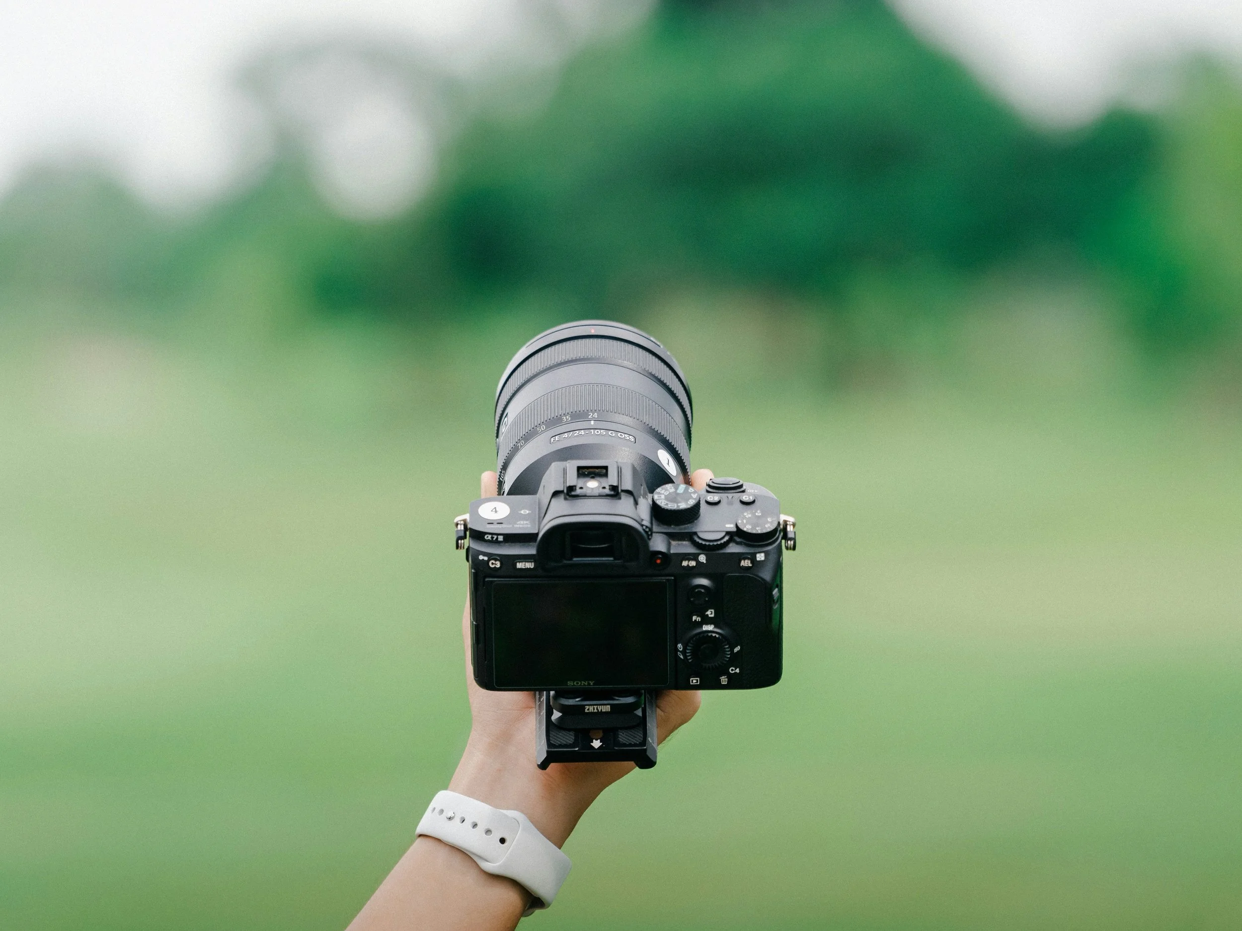 Photograph of a person holding a Sony camera with a zoom lens, with a blurred green background.
