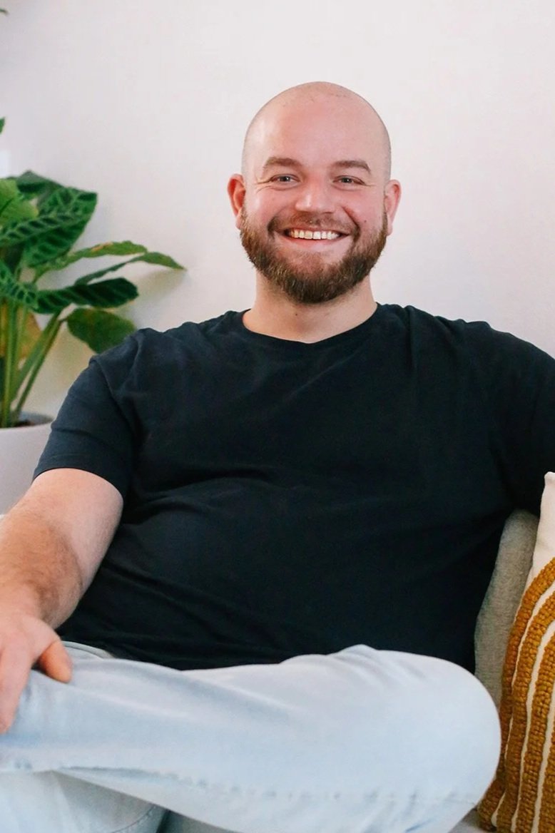 Een lachende man met een baard, zittend op een bank, met een groene plant op de achtergrond.