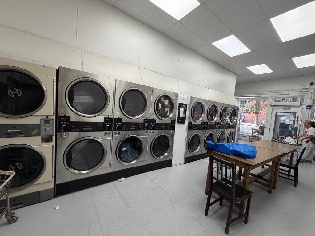 Laundry facility with multiple front-loading washing machines and dryers, wooden table with a blue bag, and a person in the background.
