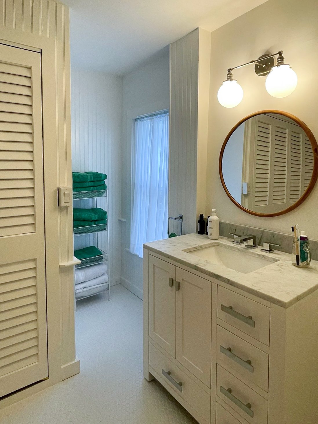 Bathroom with white cabinetry and marble countertop, round mirror, and soft lighting. Towels on a storage rack and various toiletries on the counter.