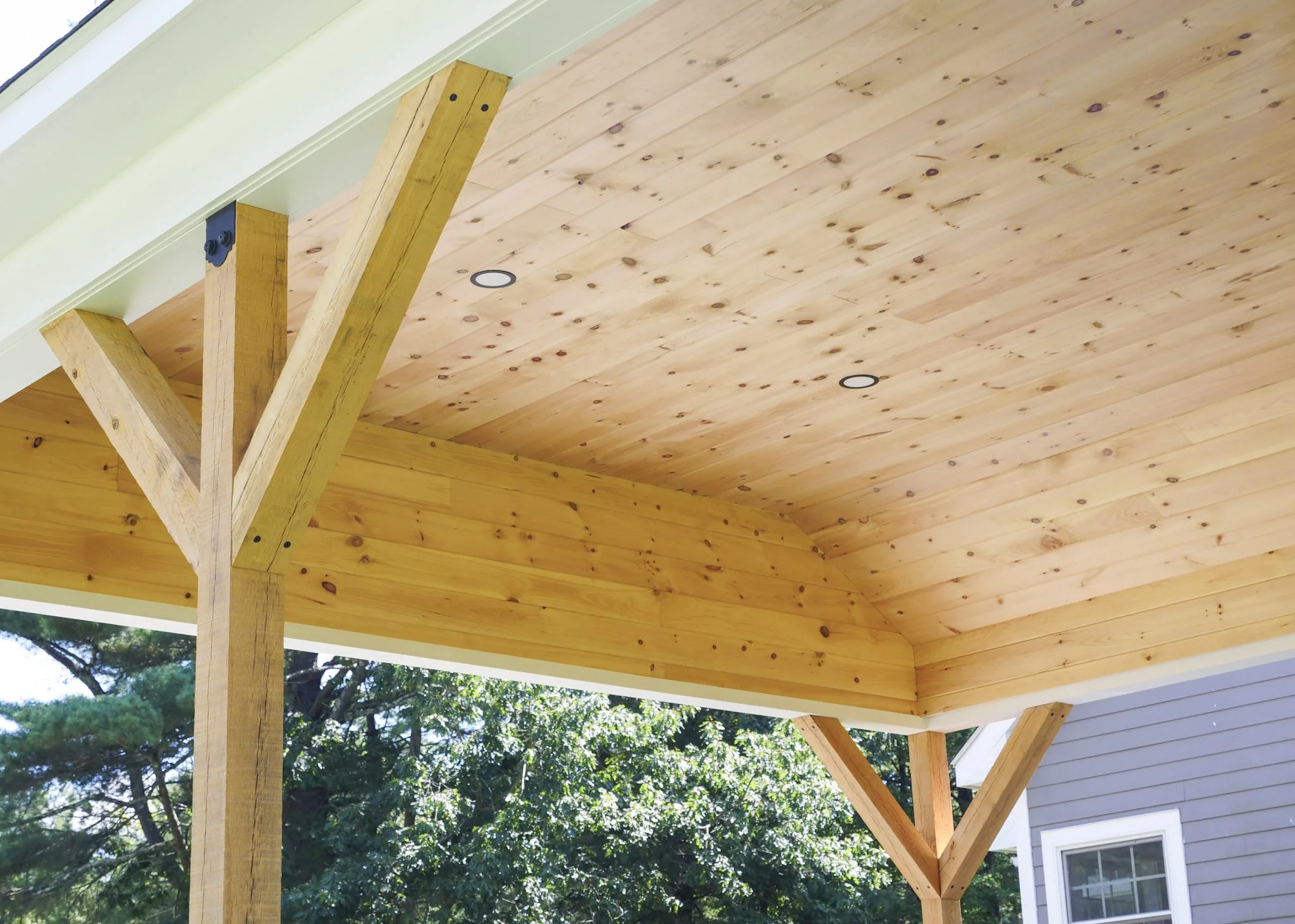 The image shows the underside of a wooden carport roof with a natural wood finish, supported by wooden beams, and the house exterior with a window and siding visible in the background. There are trees and greenery outside.