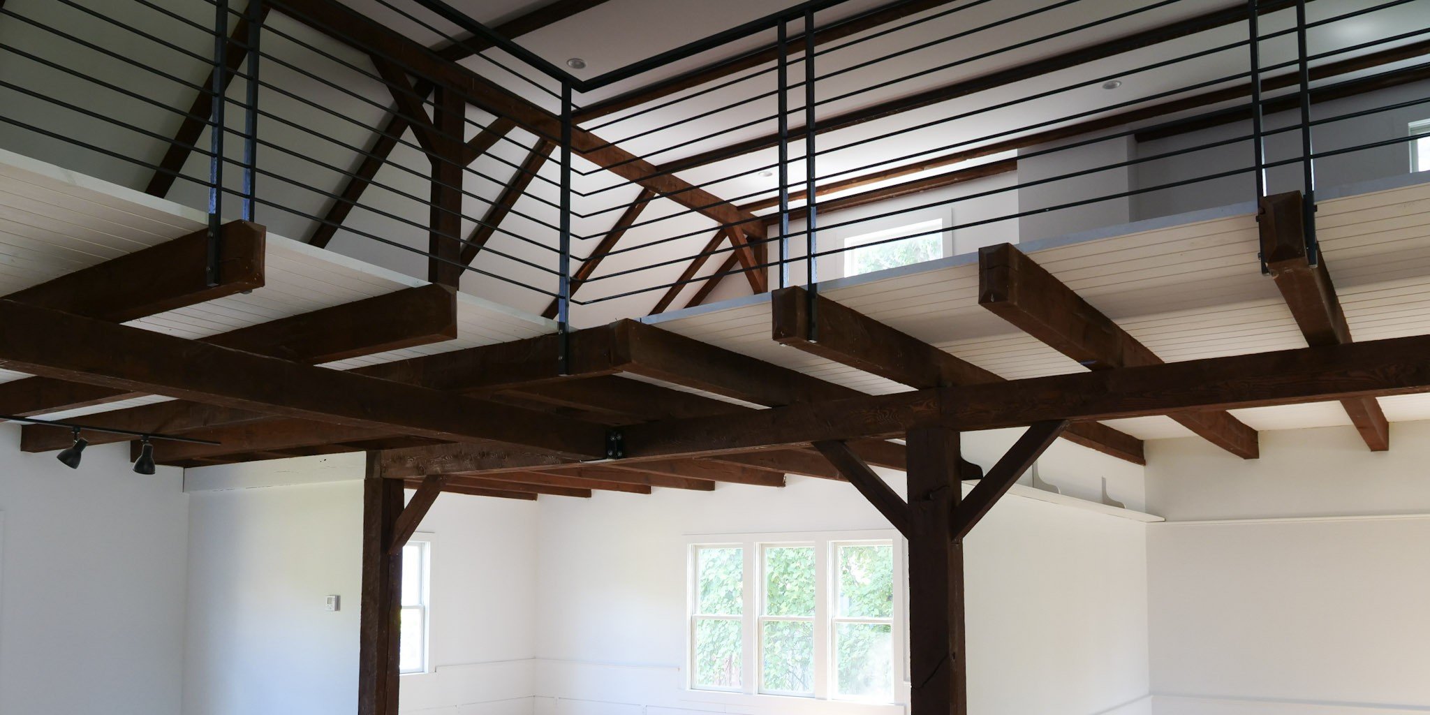 Interior view of a room with a wooden loft or mezzanine level supported by large wooden beams and black metal railings, with windows letting in natural light.